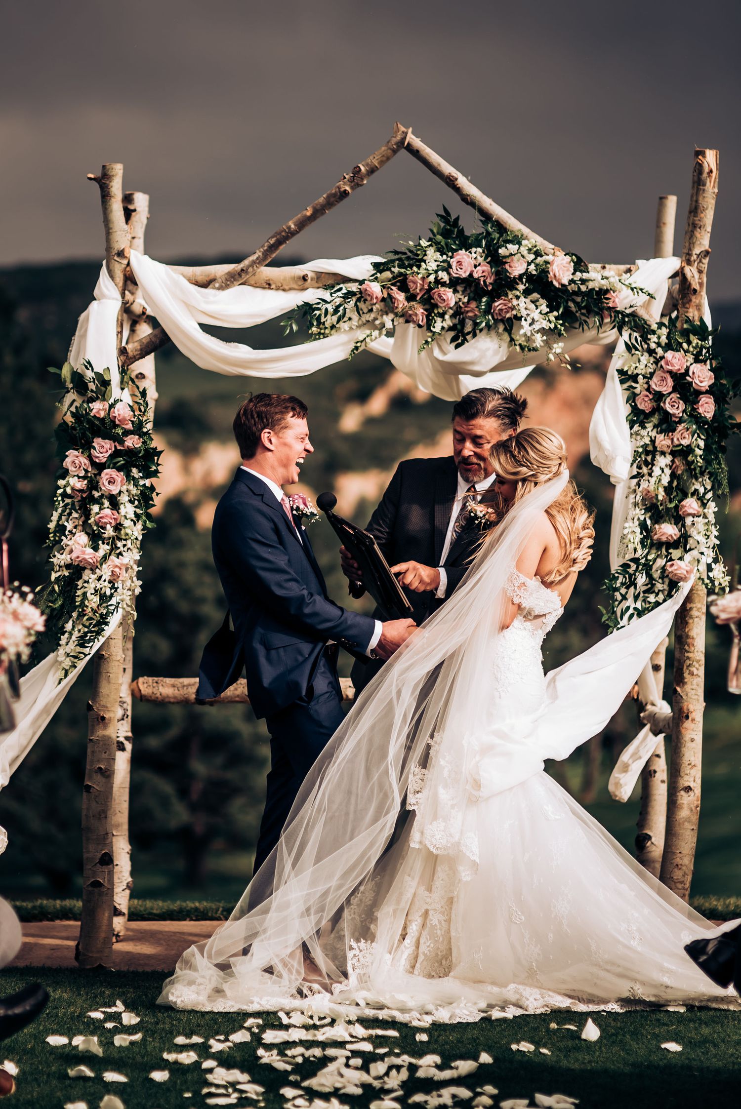 Wedding ceremony under rustic birch branch arch decorated with white fabric and pink flowers.