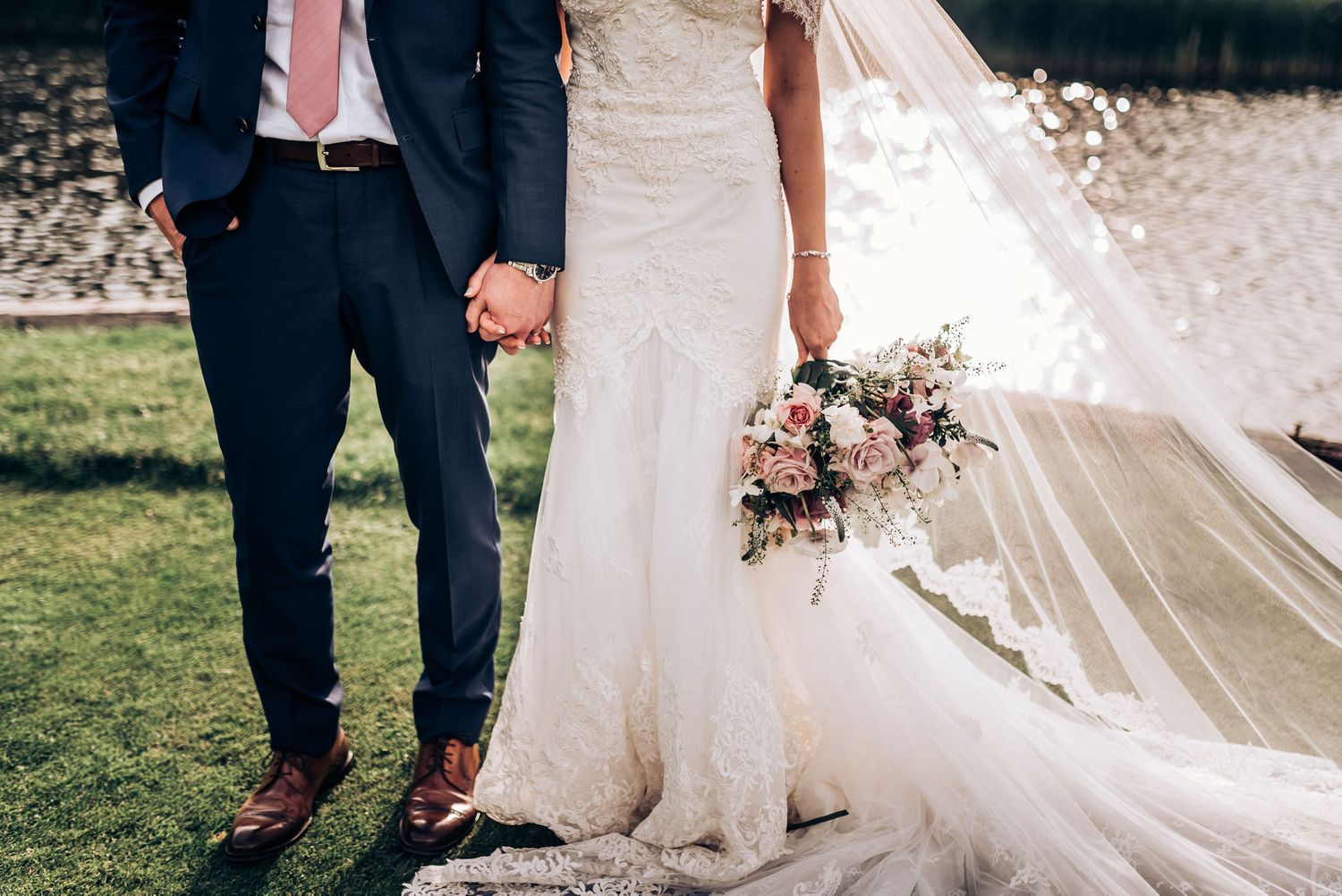 Detail shot of wedding couple holding hands with lace dress, navy suit and pink floral bouquet.