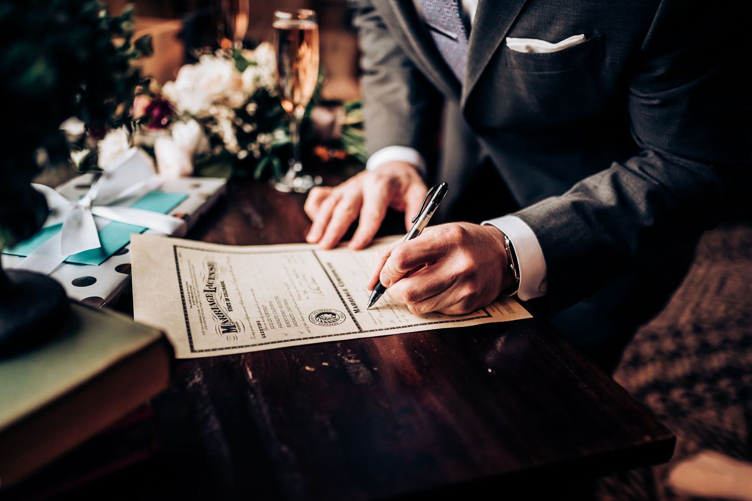 Close-up of hands signing marriage certificate with pen on dark wooden surface.