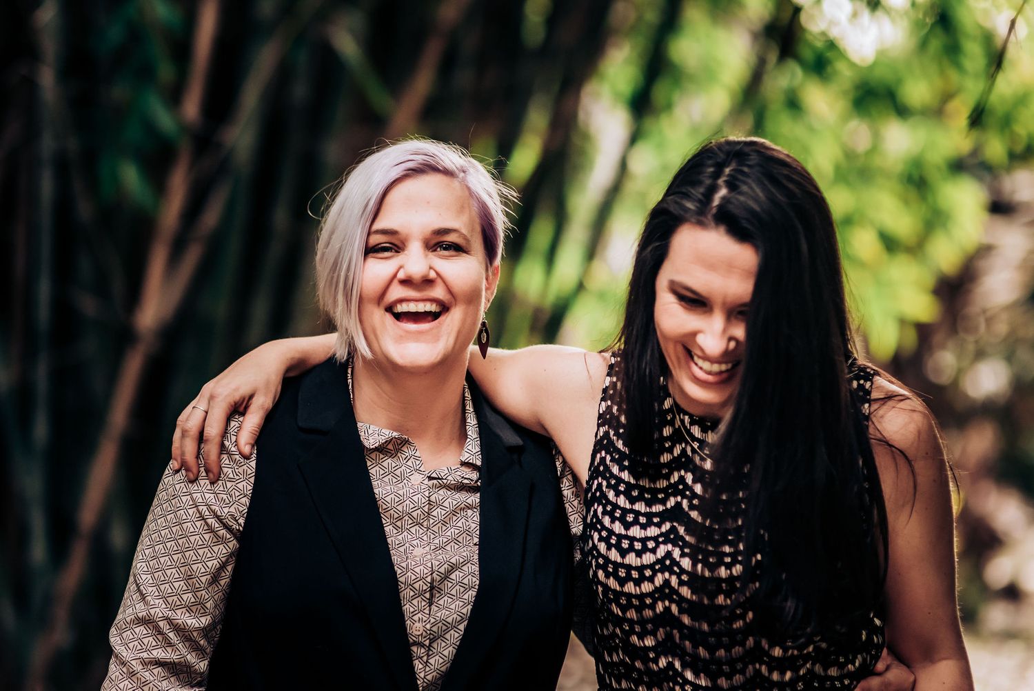 Two people share a candid laughing moment outdoors against a leafy backdrop.