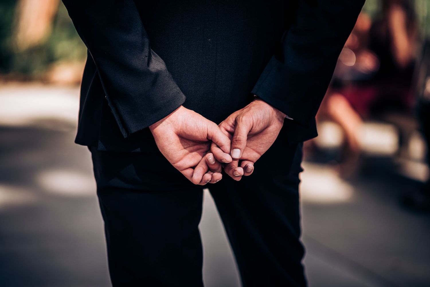 Close-up of hands clasped behind back wearing formal black suit at outdoor event.