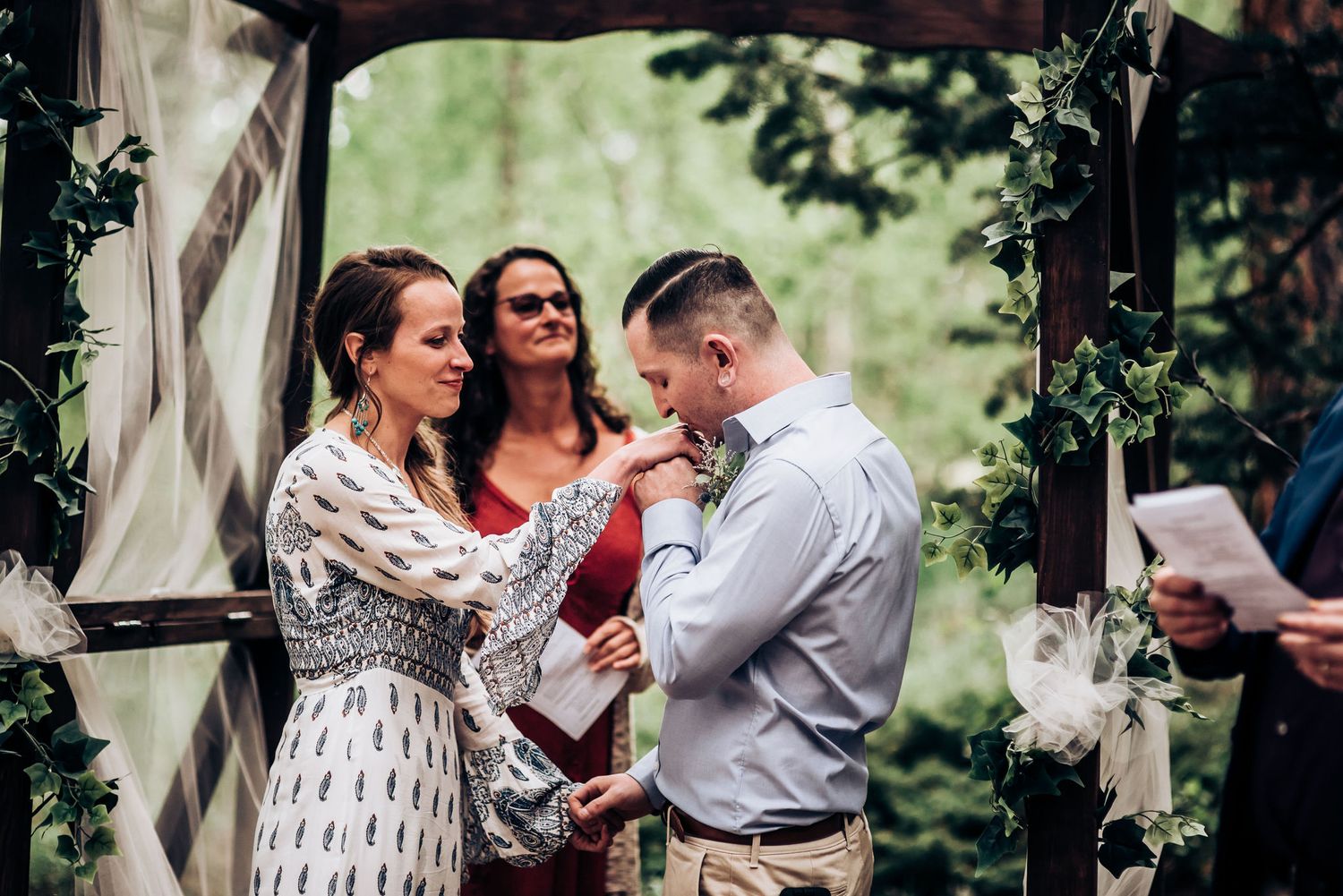 A wedding ceremony takes place under a rustic floral arch in a forest setting with an officiant leading the service.