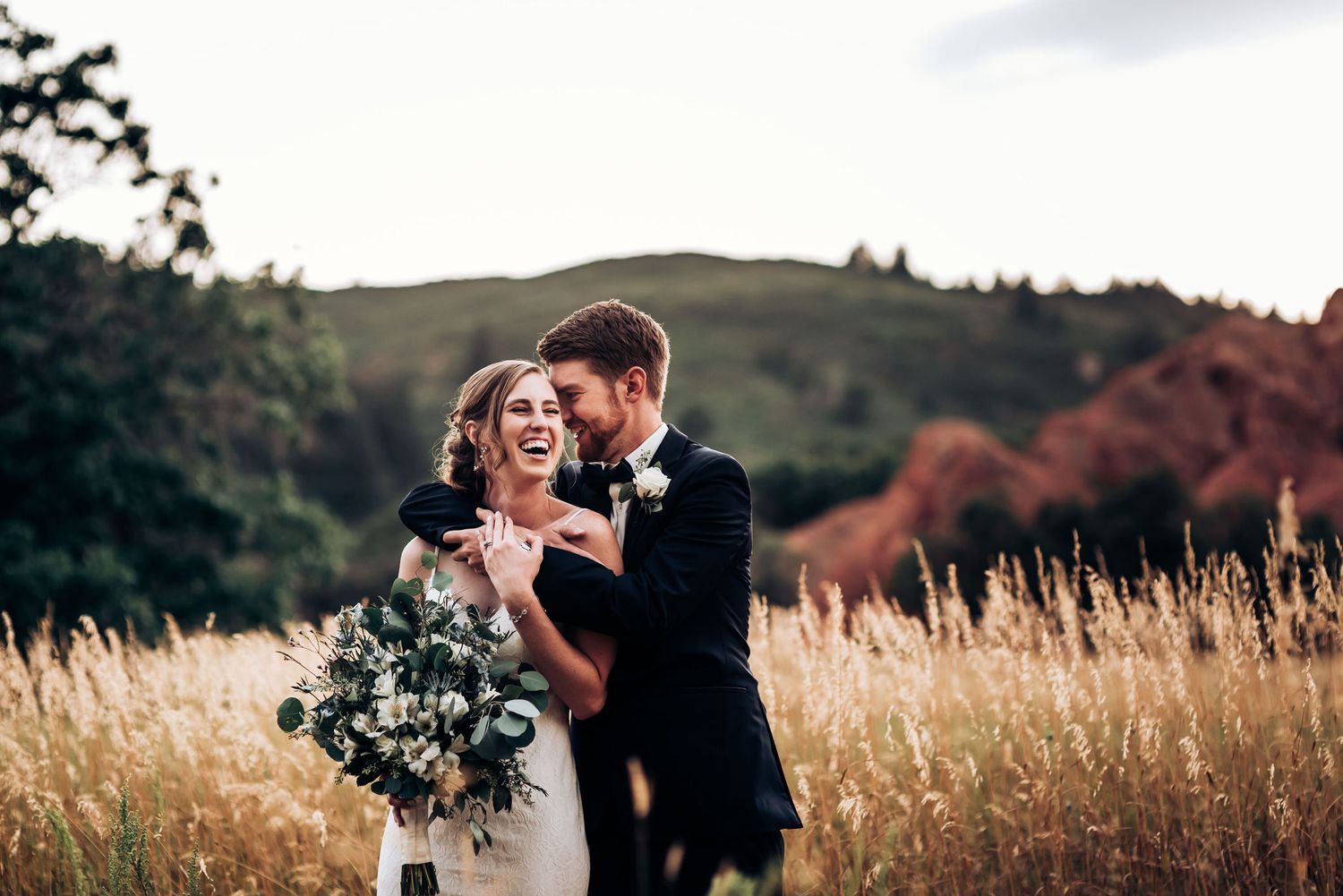 Wedding couple laughs together in rustic field during sunset photoshoot.