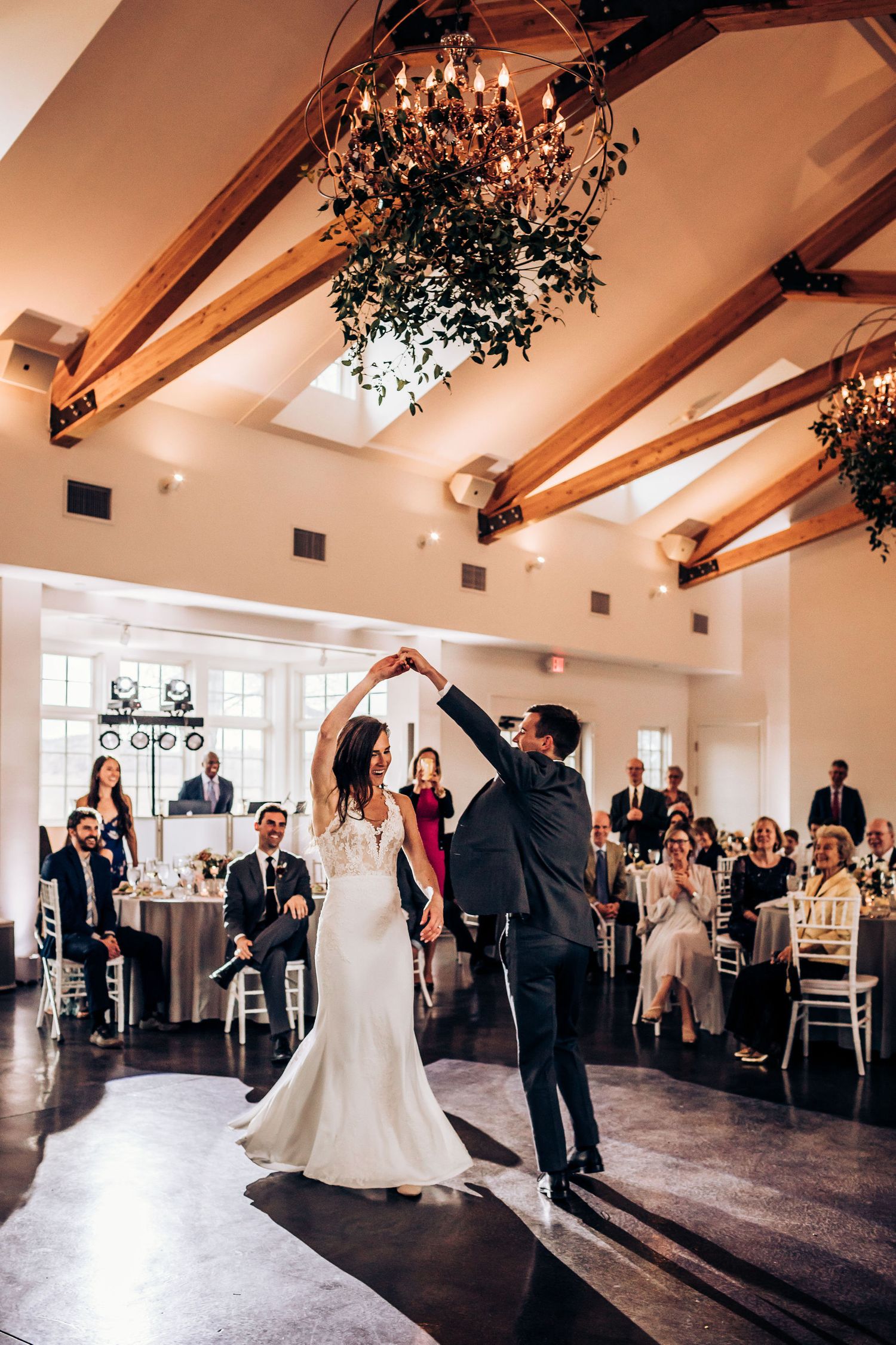 A couple performs their first dance under a chandelier in a rustic venue with exposed wooden beams.