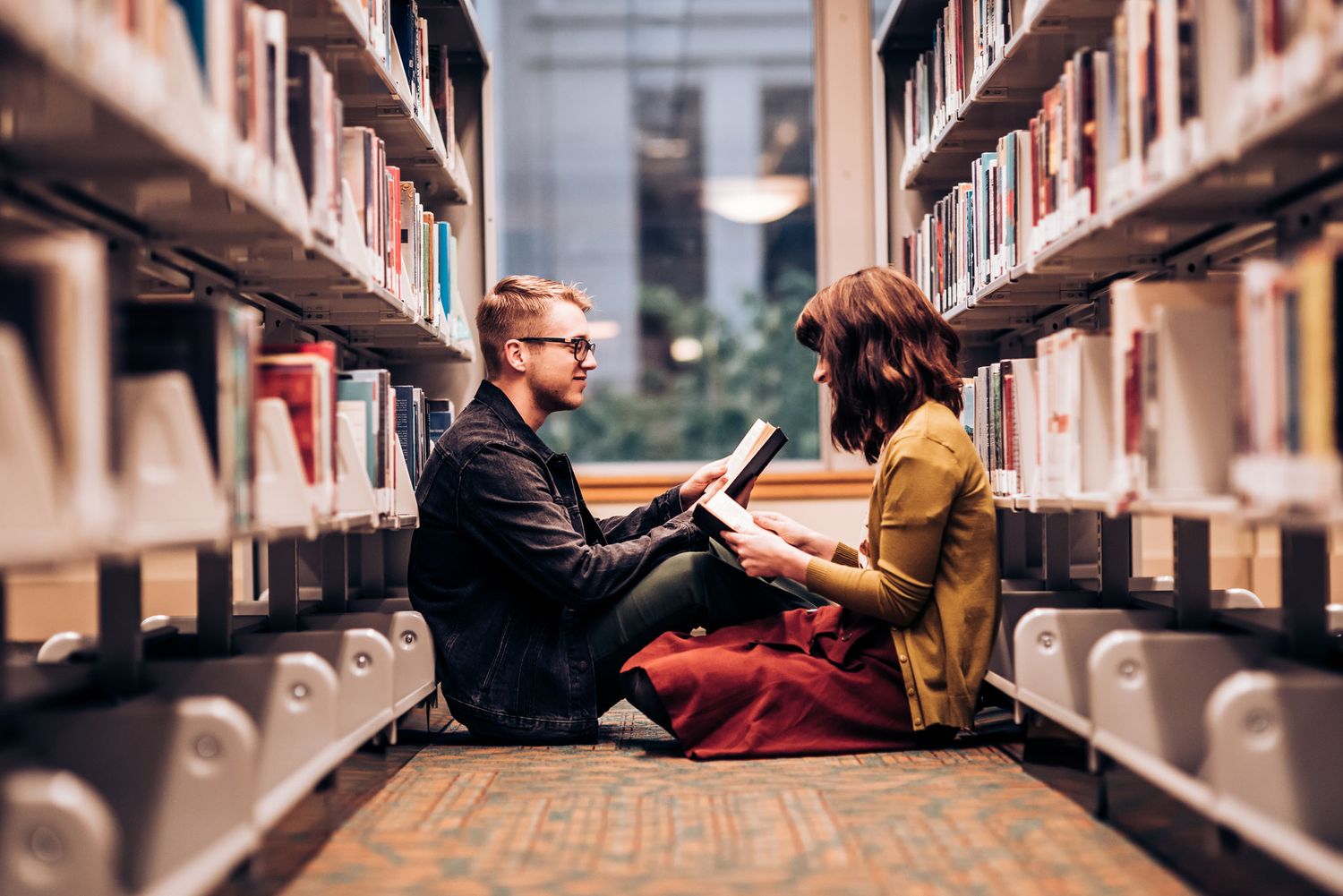 A couple sits together between rows of library bookshelves.