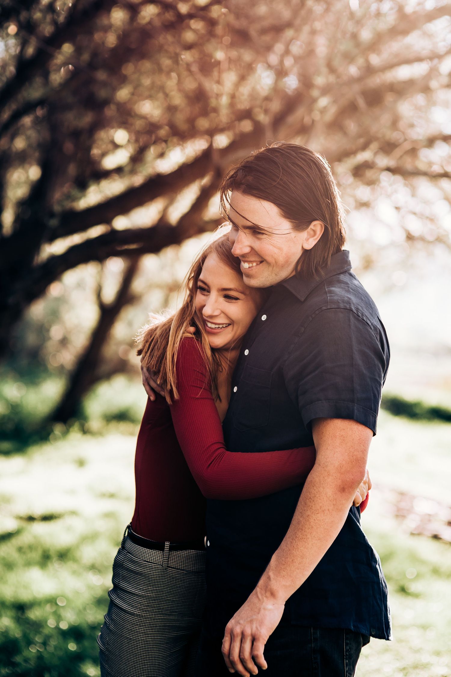A romantic couple embraces and laughs together in dappled sunlight under autumn trees.