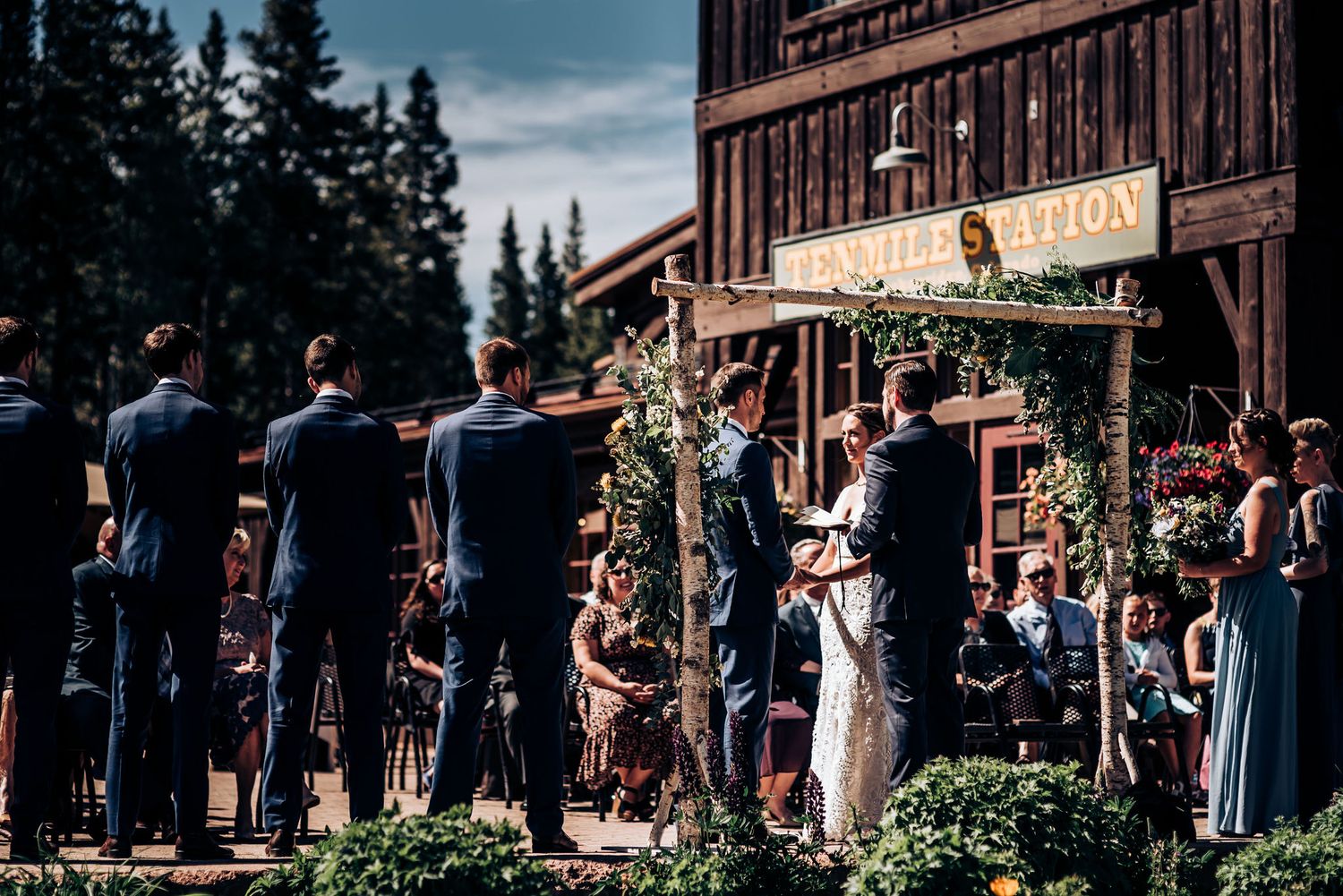 Wedding party stands at rustic venue with wooden building and floral decorations.