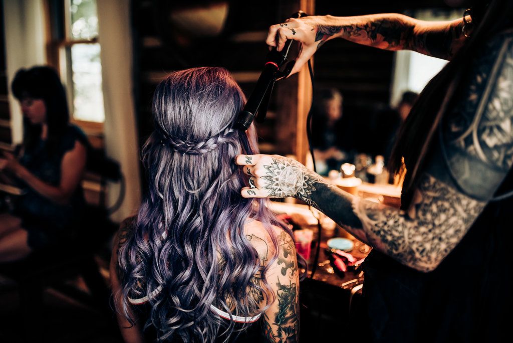 A hairstylist works on long wavy purple-tinted hair in a dimly lit room.