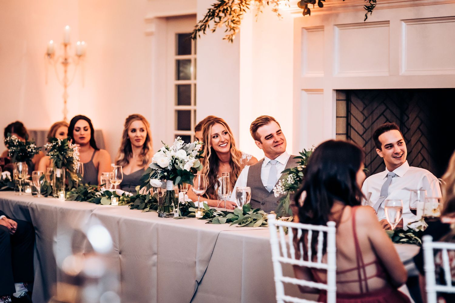 Wedding party seated at long head table decorated with greenery and soft lighting.