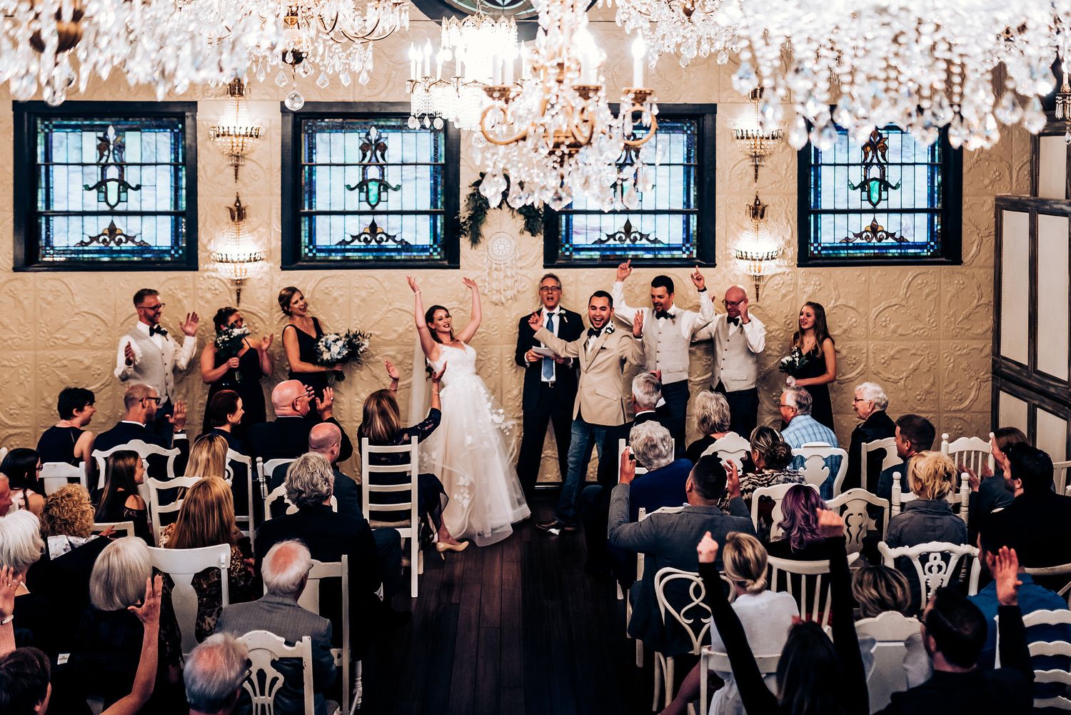 Wedding ceremony in a venue with crystal chandeliers and stained glass windows along textured walls.