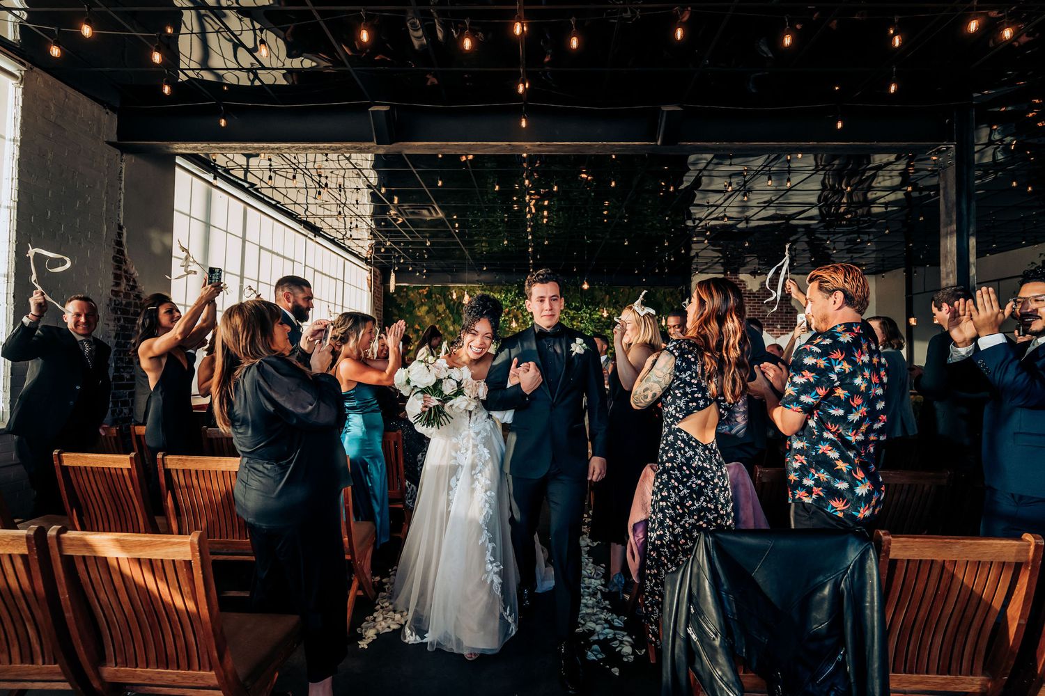 Wedding guests celebrate and cheer during a reception in an industrial-style venue with exposed ceiling and string lights.