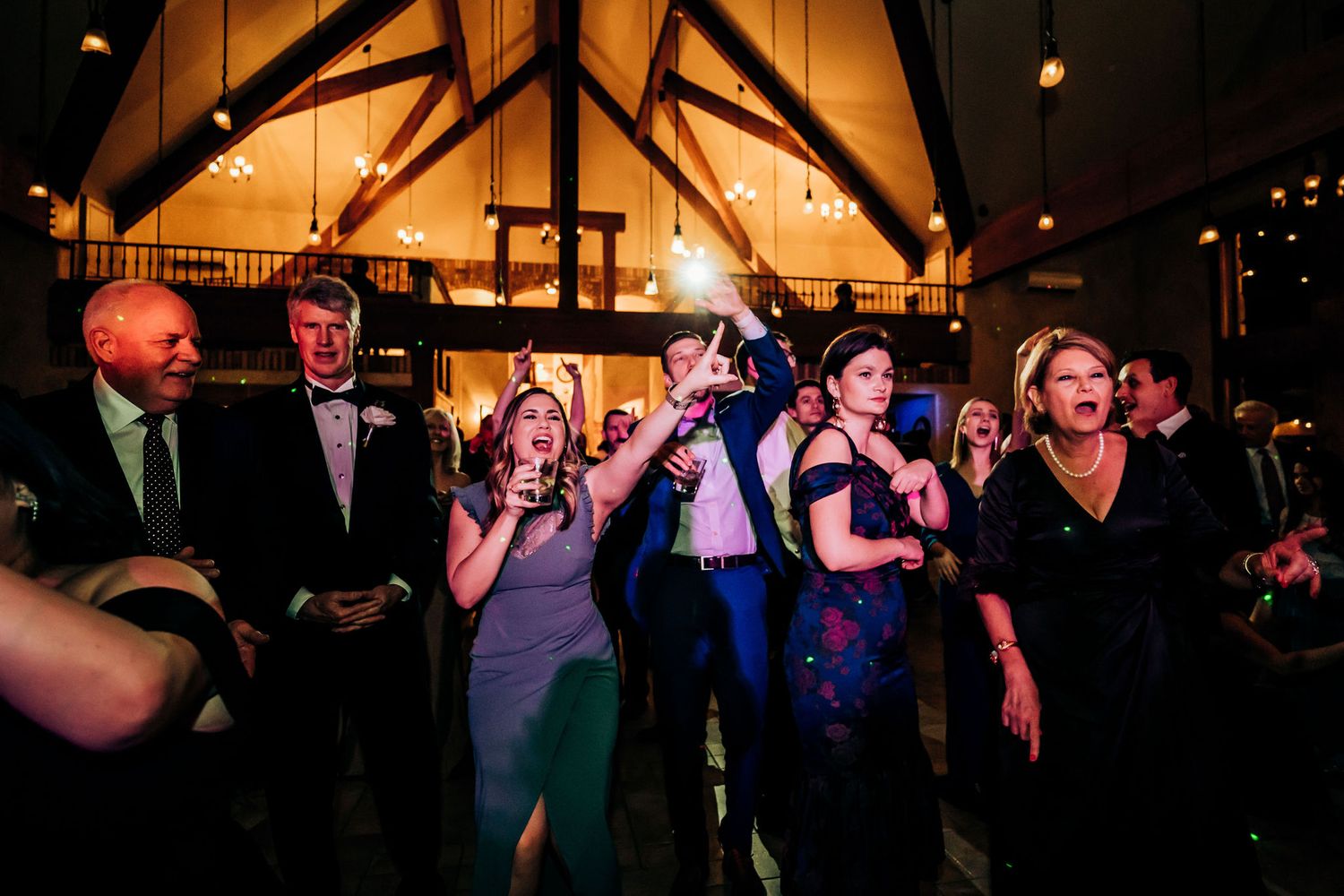 Party guests dancing and celebrating under wooden beam ceiling with festive lighting.