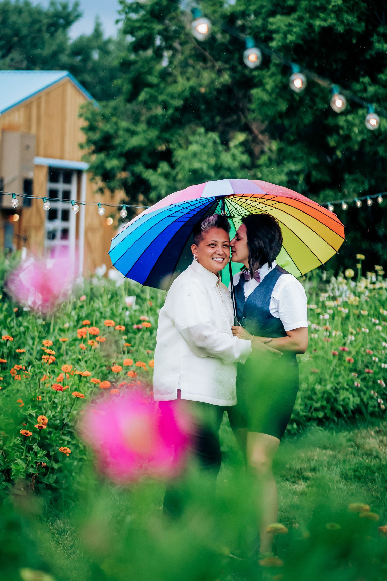 Two people share a rainbow umbrella in a garden with string lights and pink flowers.