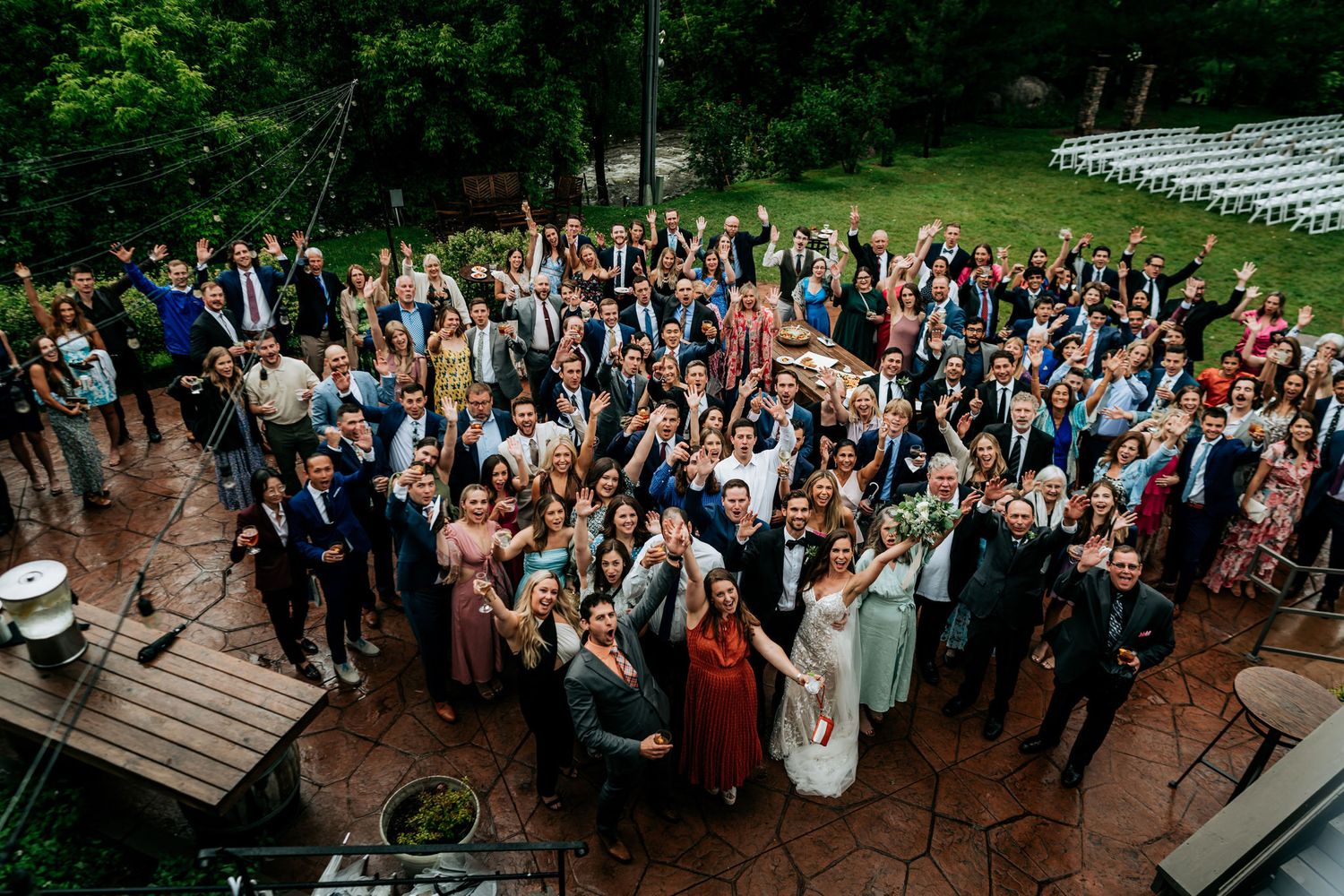 Large crowd of wedding guests gathered outdoors for a group photo on wooden deck.