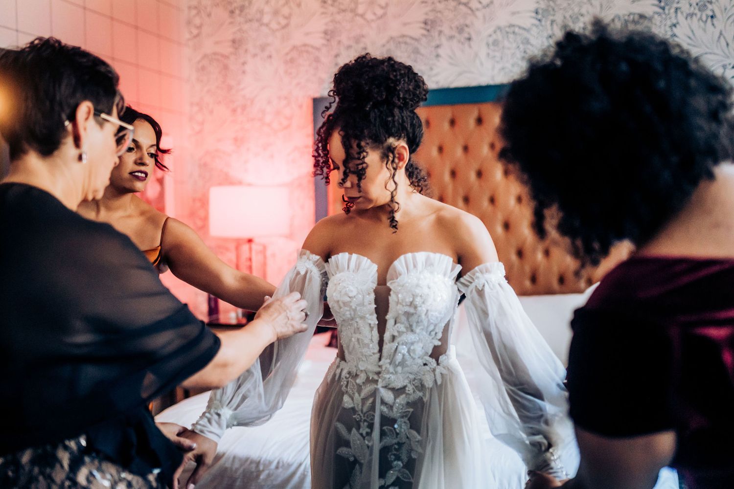 A bride in an ornate white gown gets final adjustments while surrounded by helpers in a decorated room.