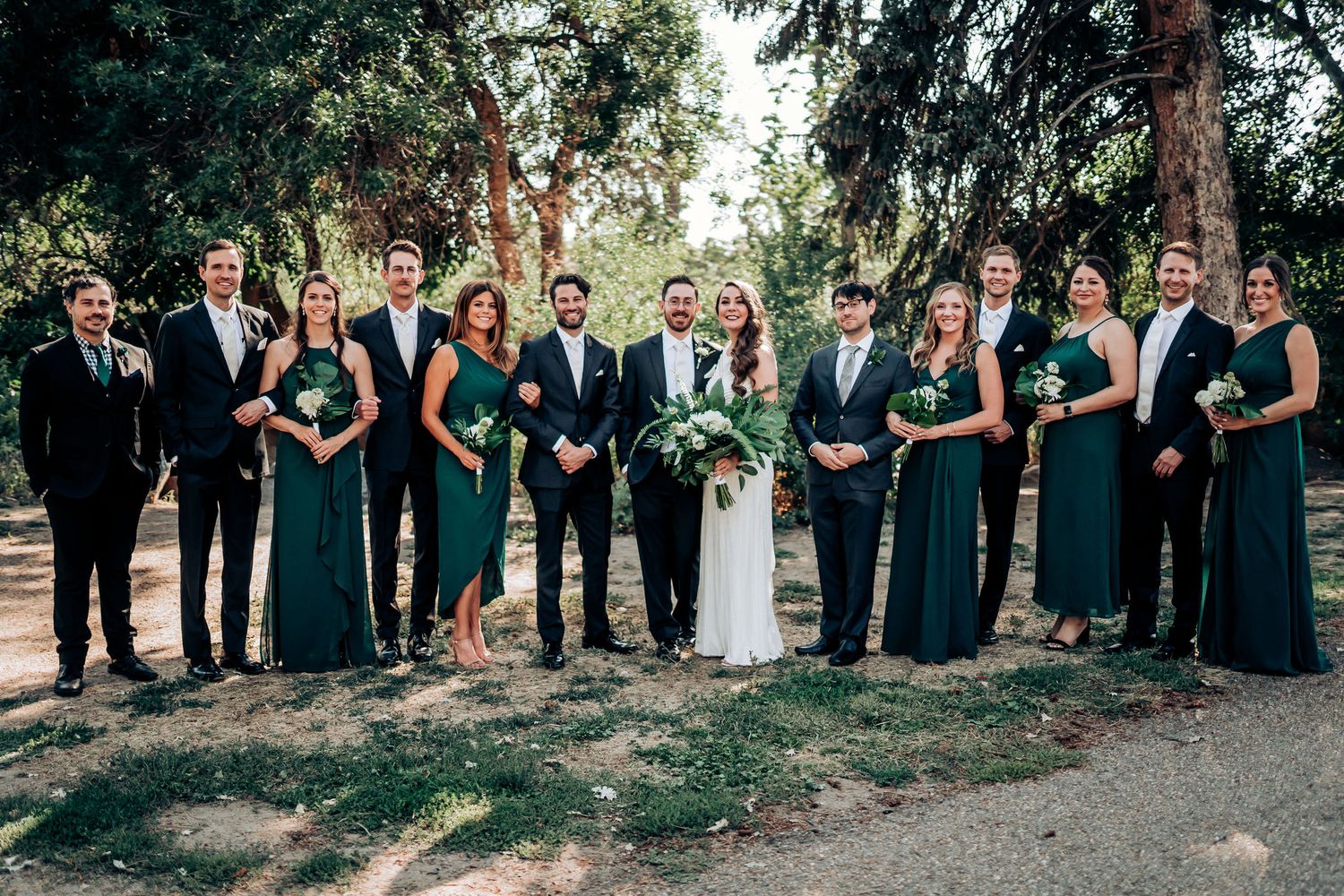 Large wedding party in emerald and black attire gathered for formal group photo outdoors.