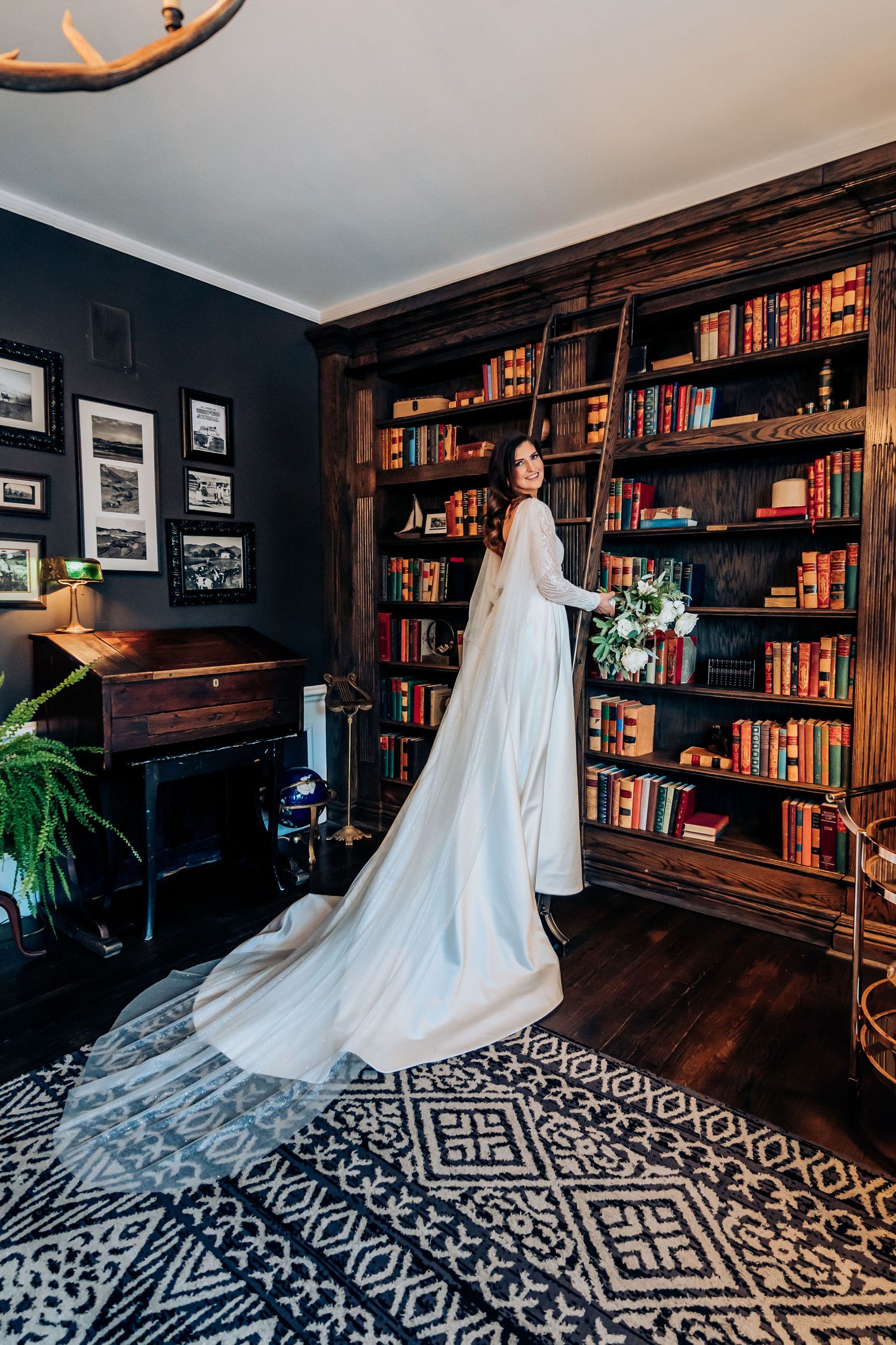 Bride's flowing train spreads across geometric rug in classic library setting.