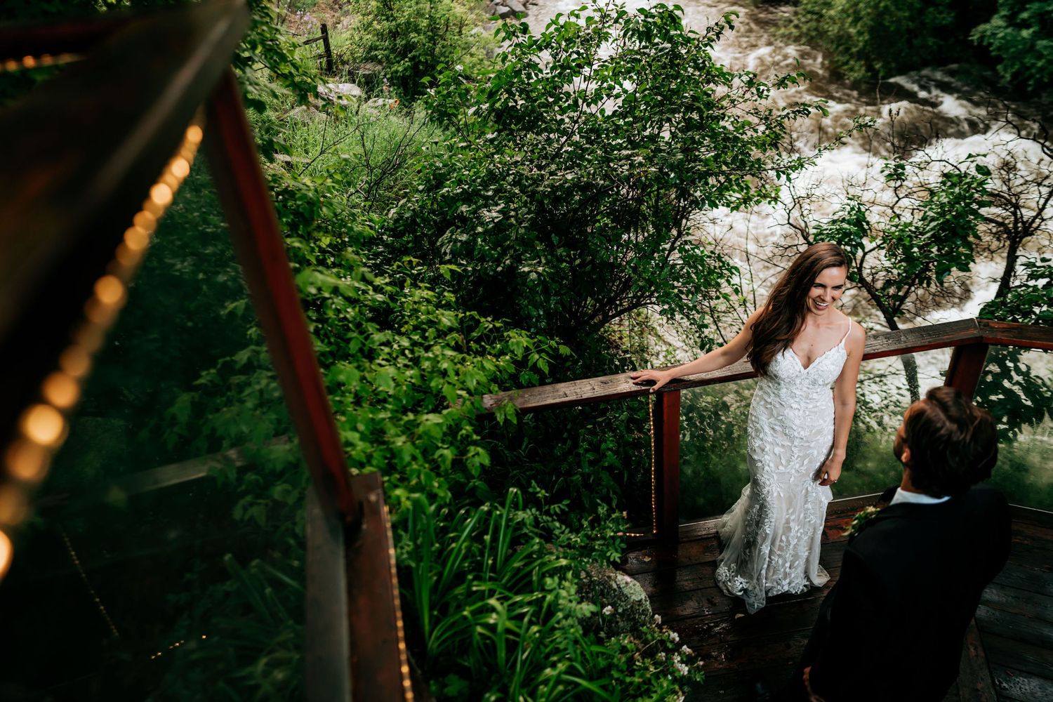A bride in a fitted lace dress stands on a rustic wooden deck surrounded by greenery.
