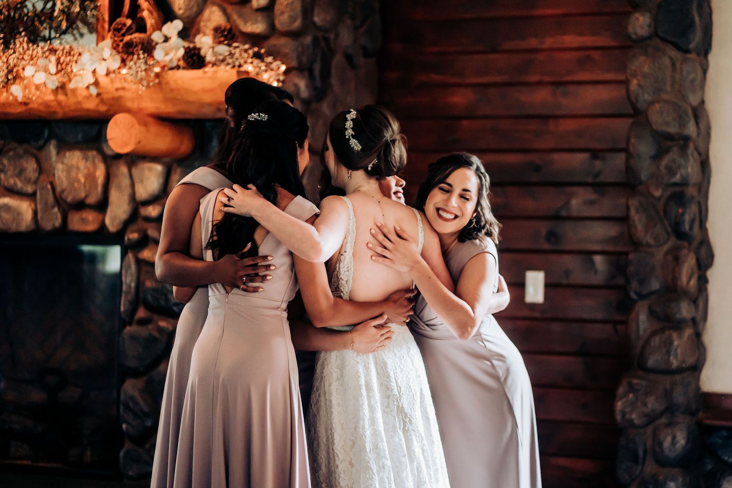 Bridesmaids in soft pink dresses share an emotional embrace with the bride at a rustic venue.