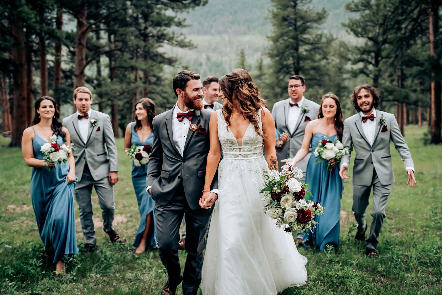 Bridal party walks together through mountain forest meadow wearing blue dresses and gray suits.