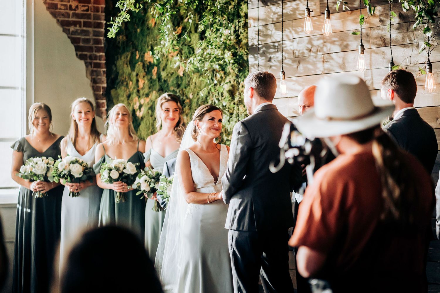 Wedding ceremony in a rustic brick venue with hanging greenery and attendants in dark gowns.