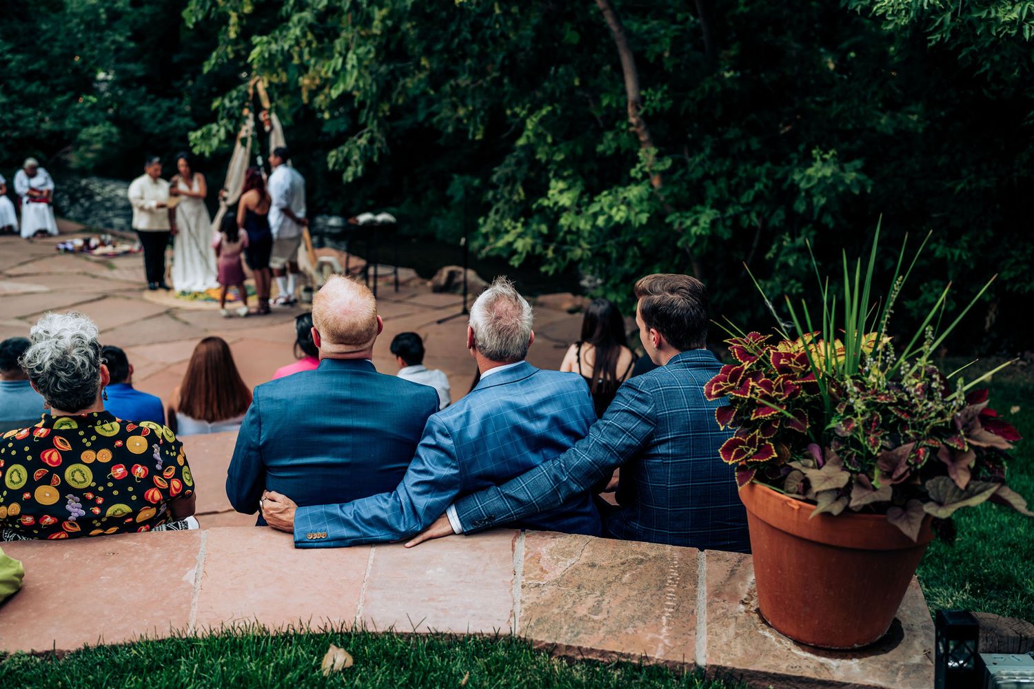 Guests seated on stone wall watching outdoor wedding ceremony with decorative potted plants.