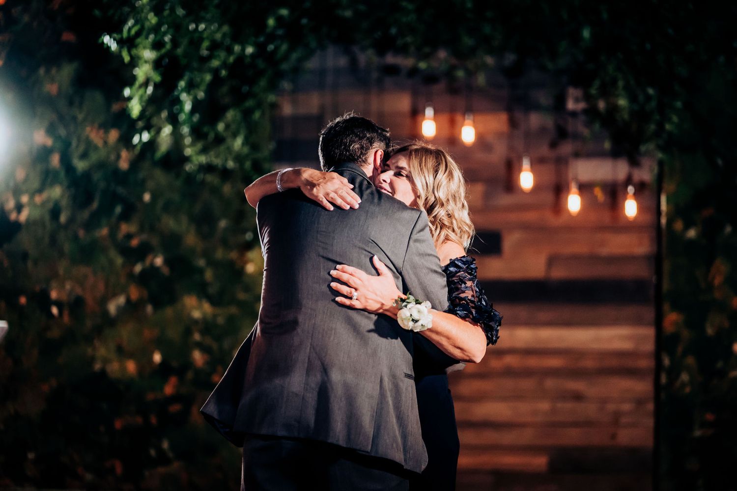 Tender embrace captured during evening wedding dance with ambient lighting and greenery.
