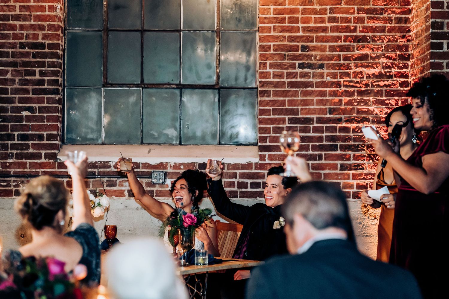 Wedding guests react enthusiastically to speeches during a reception in a brick-walled venue.