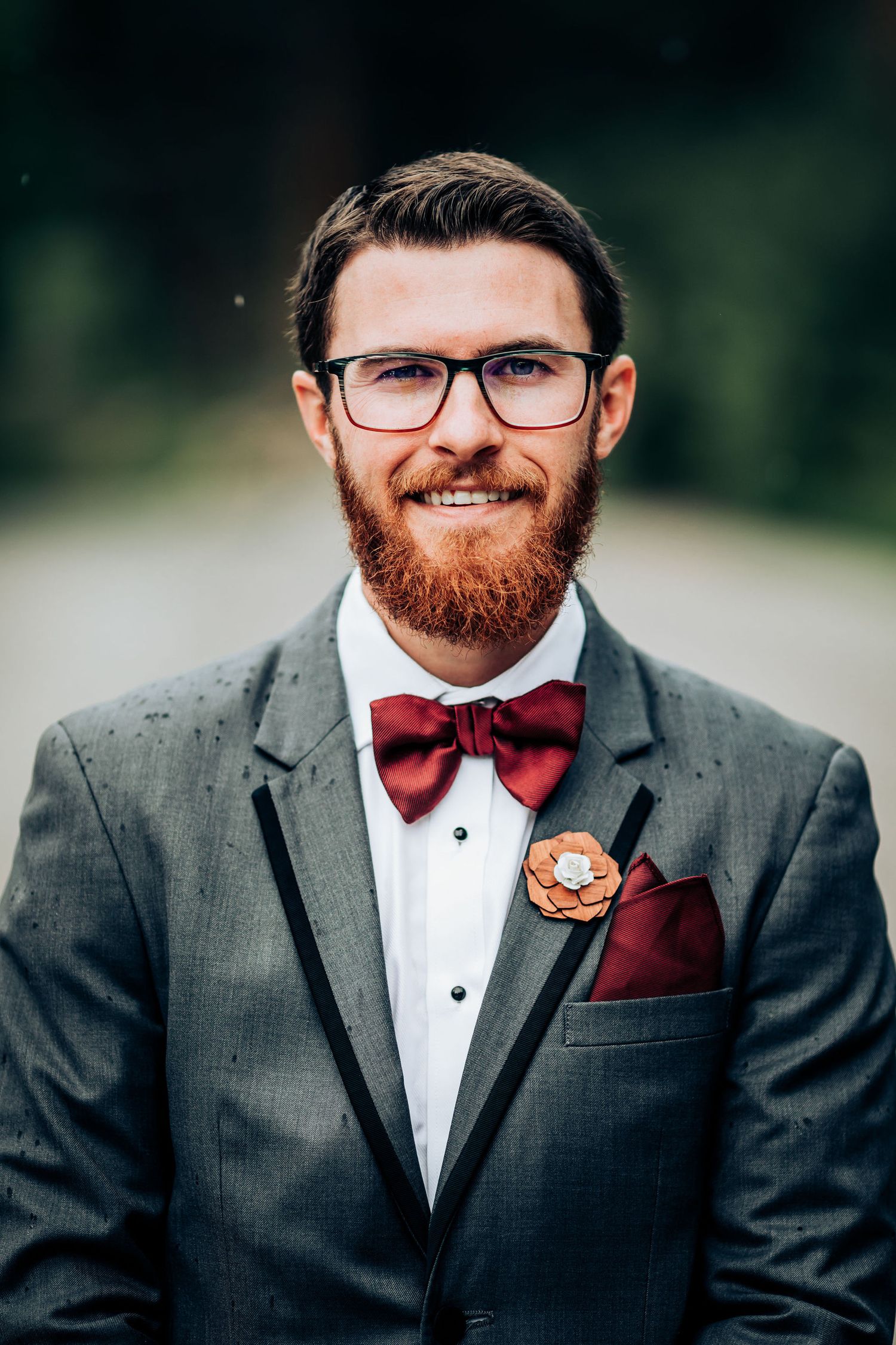A formal portrait showing wedding attire with burgundy bowtie and boutonniere against natural background.