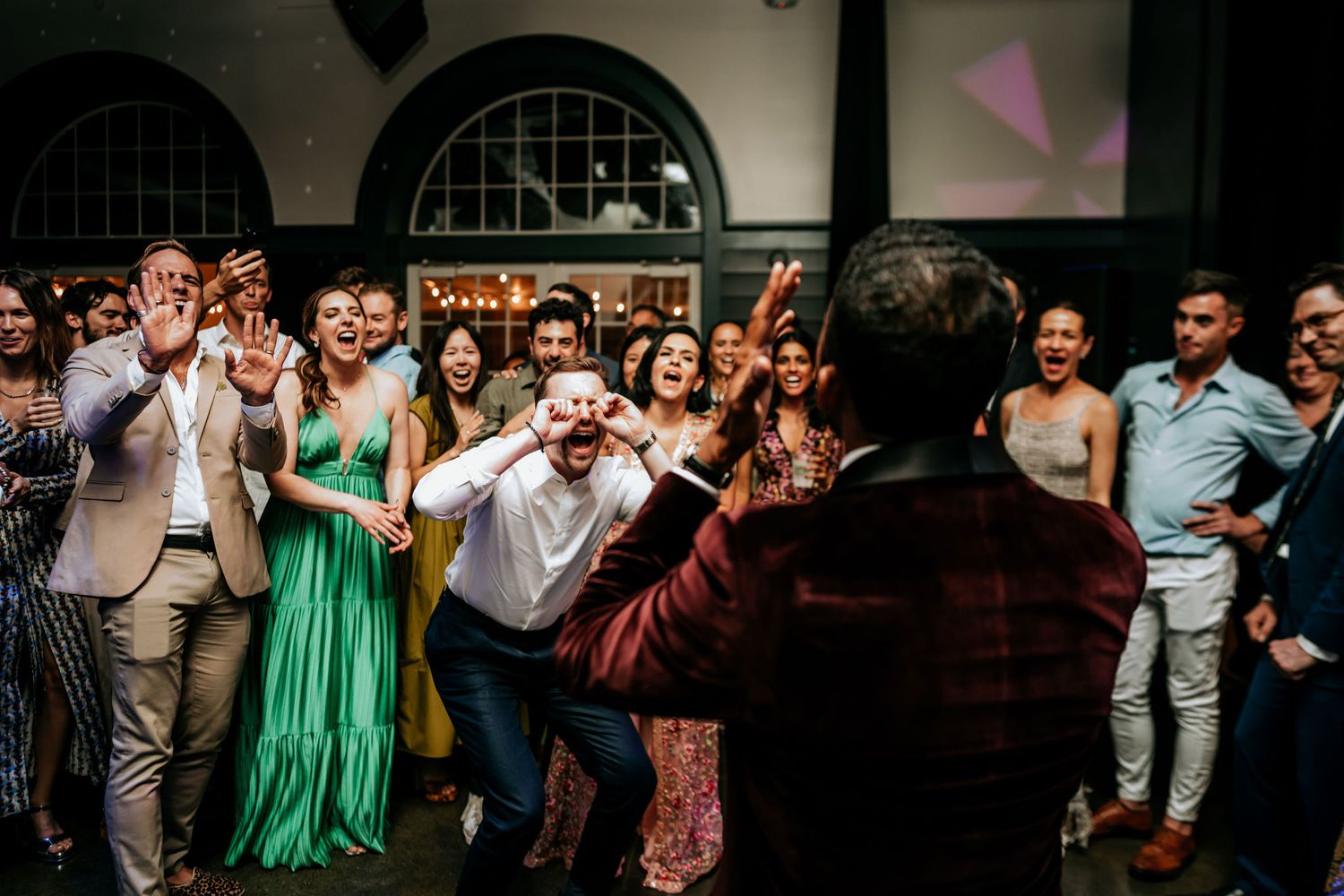 Wedding party guests dancing and celebrating in a venue with arched windows.