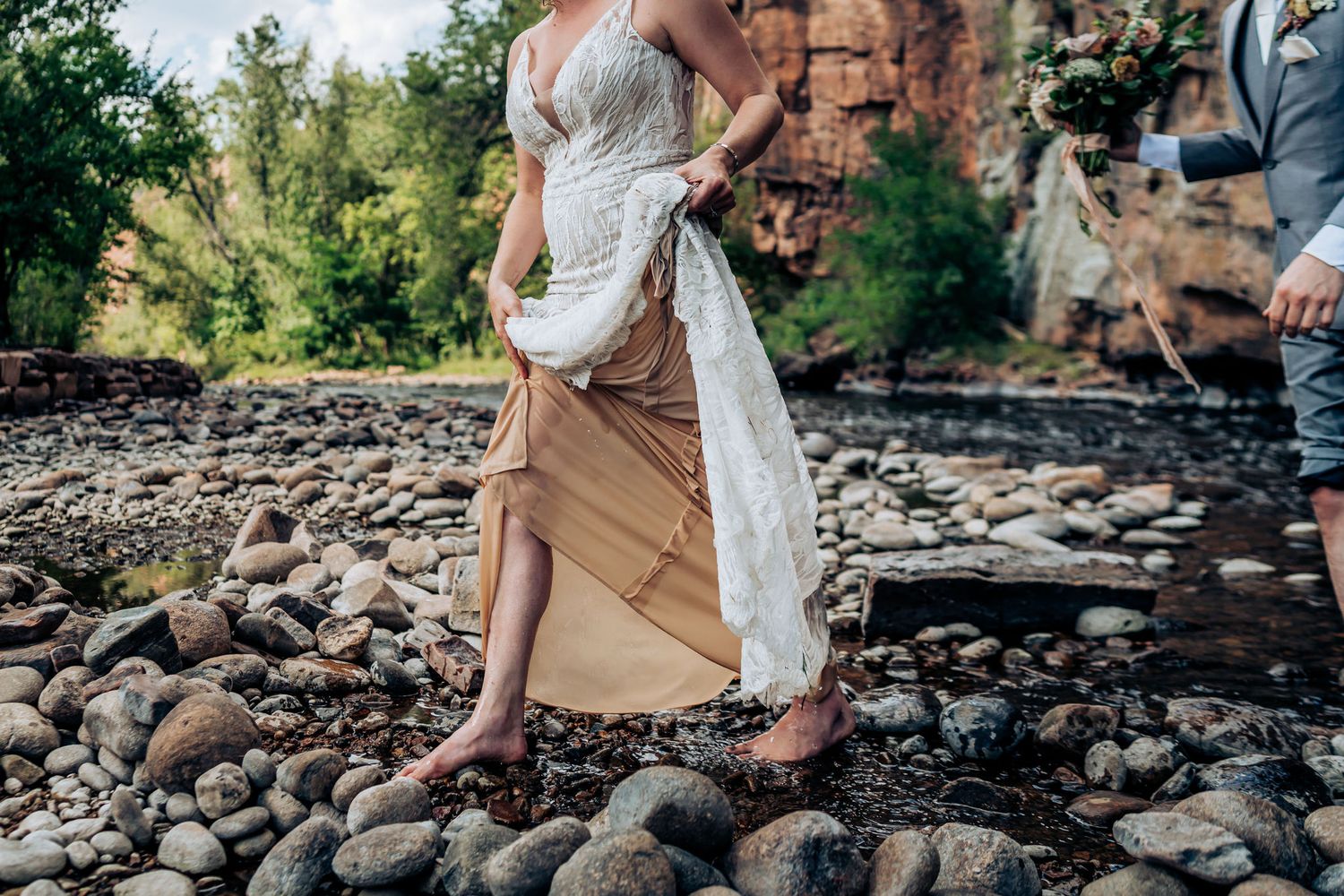 A detail shot of a bride's lace wedding dress and boots on a rocky riverbed.