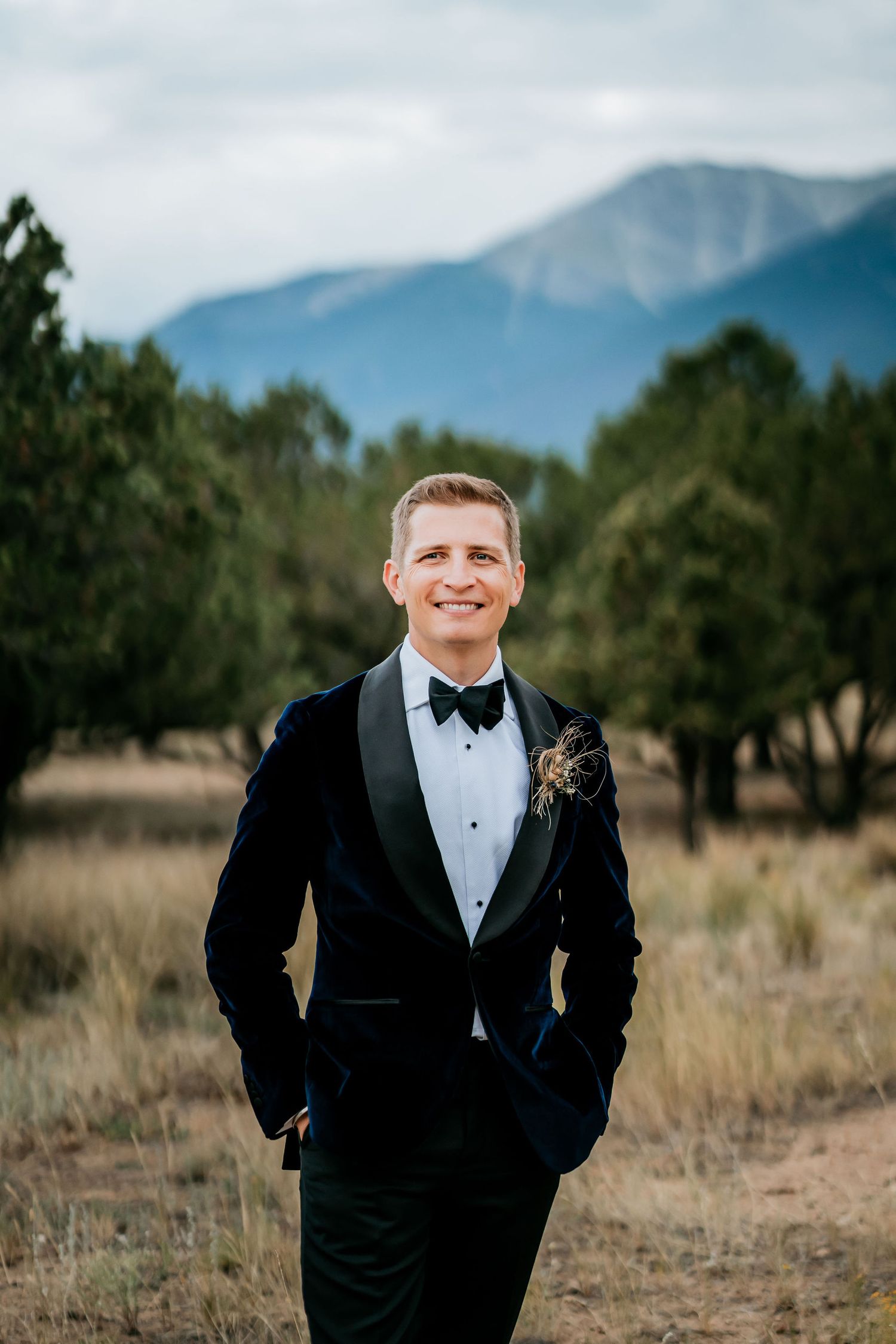 Groom in classic black tuxedo poses in front of mountain landscape with a boutonniere.
