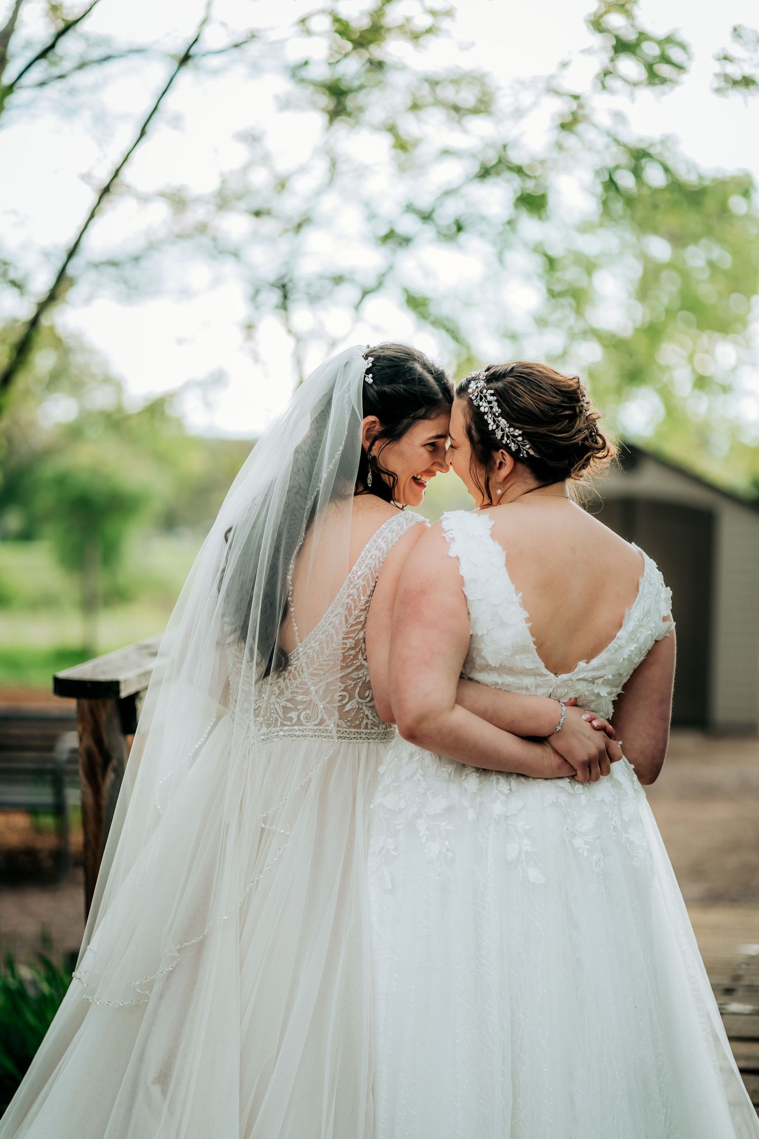 Couple in elegant white wedding dresses embracing during outdoor photoshoot.