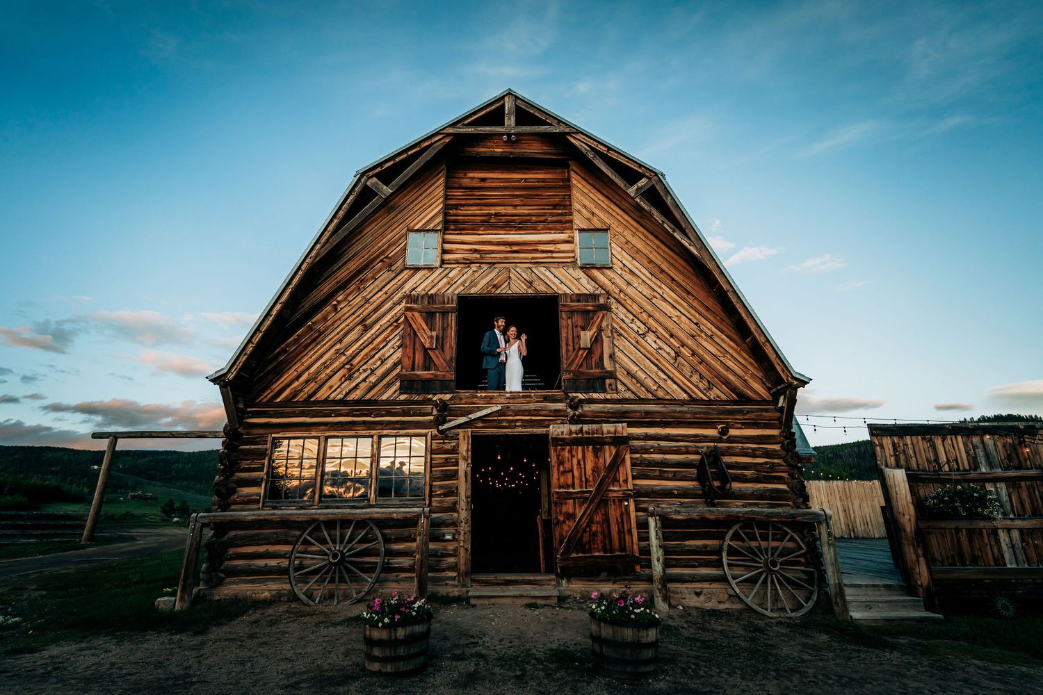 A weathered barn with vintage wagon wheels features a couple posing in the upper doorway.