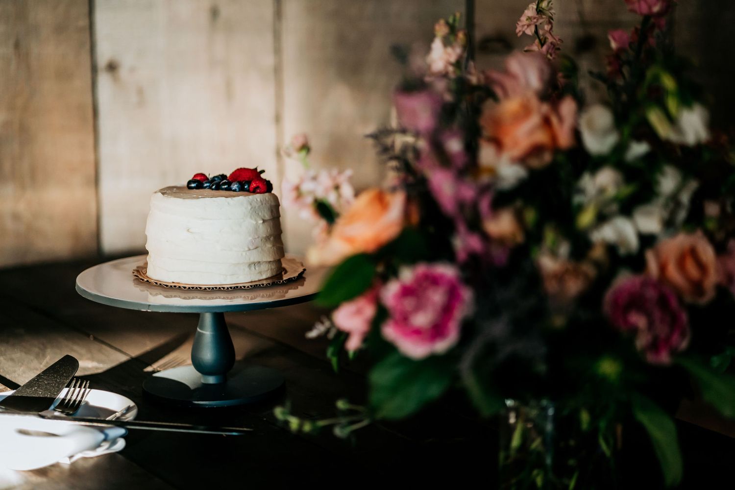 Simple white wedding cake decorated with fresh berries on dark wooden table with floral arrangement.