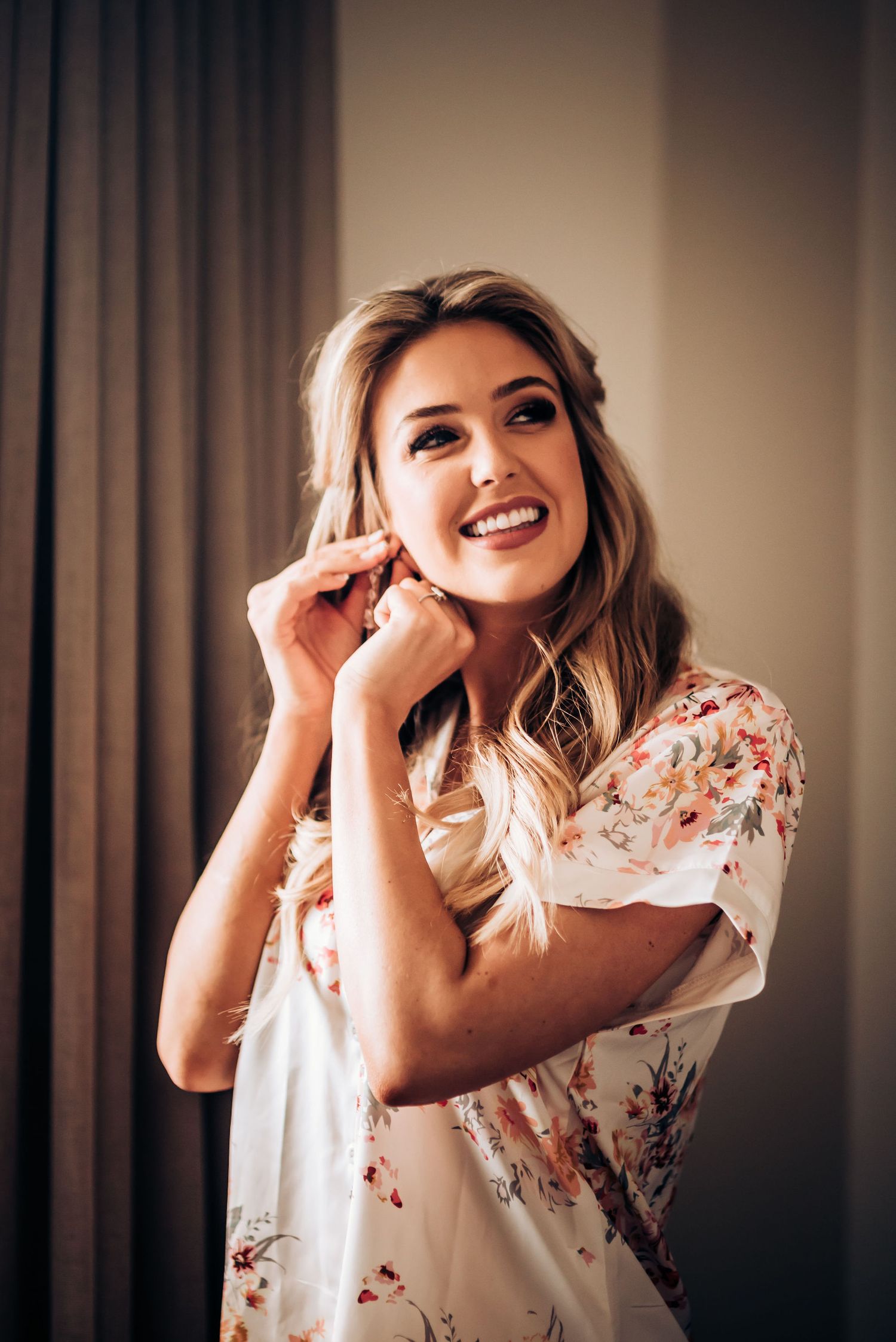 Woman in floral robe laughing while putting on earrings.