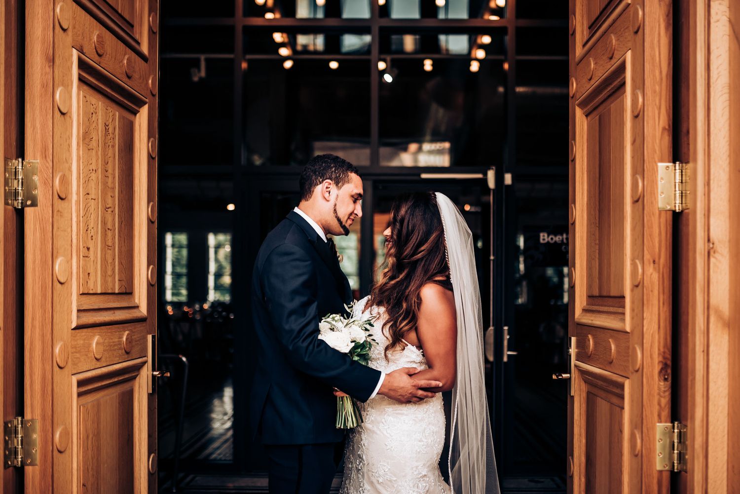 Newlyweds share an intimate moment in a doorway with elegant wooden doors and a white bridal bouquet.