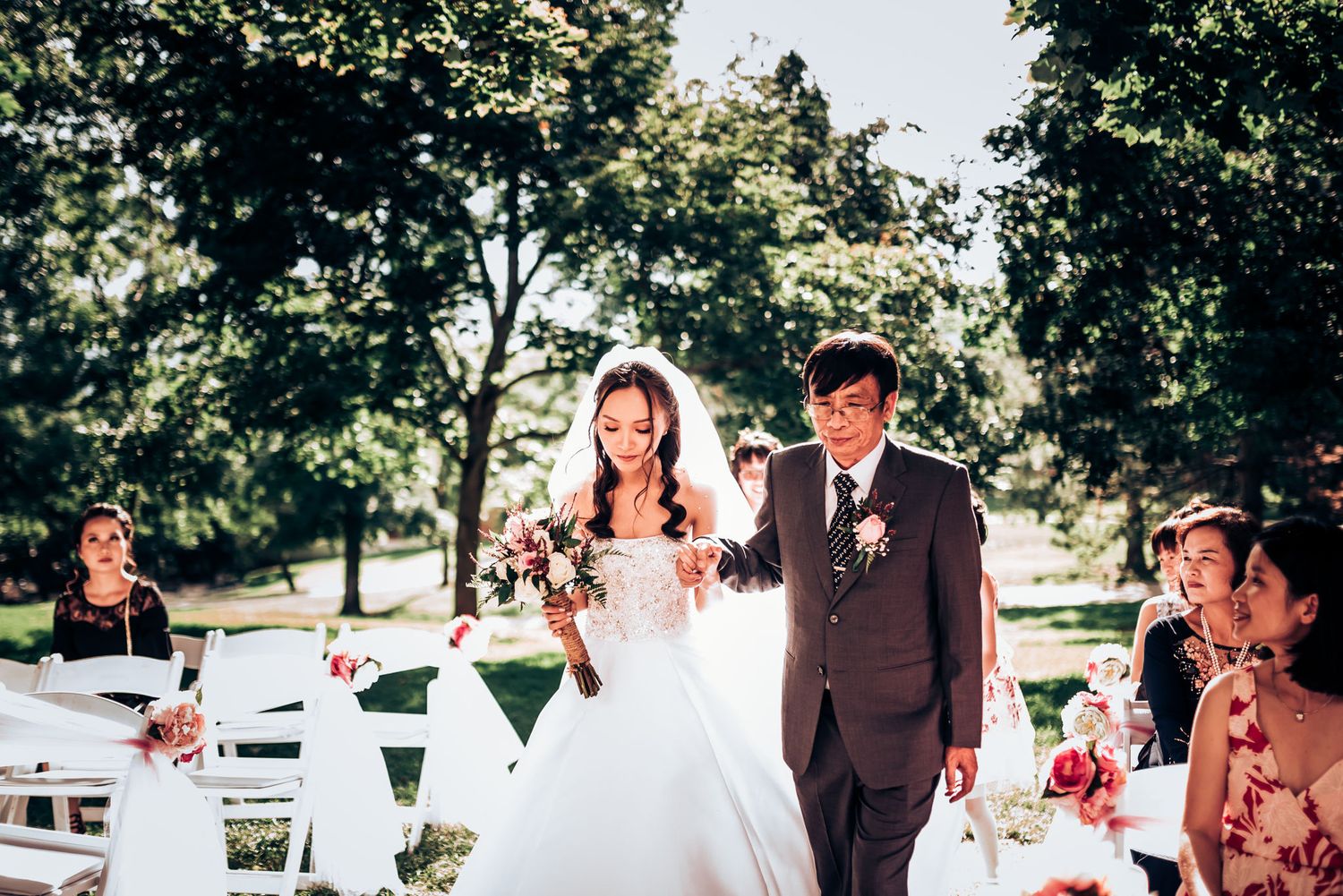 Couple walks down the outdoor wedding aisle surrounded by white chairs and trees.
