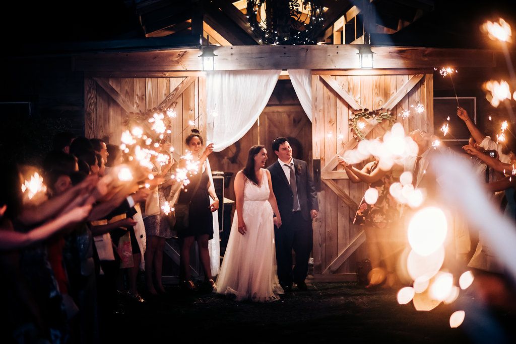 Wedding couple exits rustic barn venue while guests create magical sparkler exit tunnel at night.