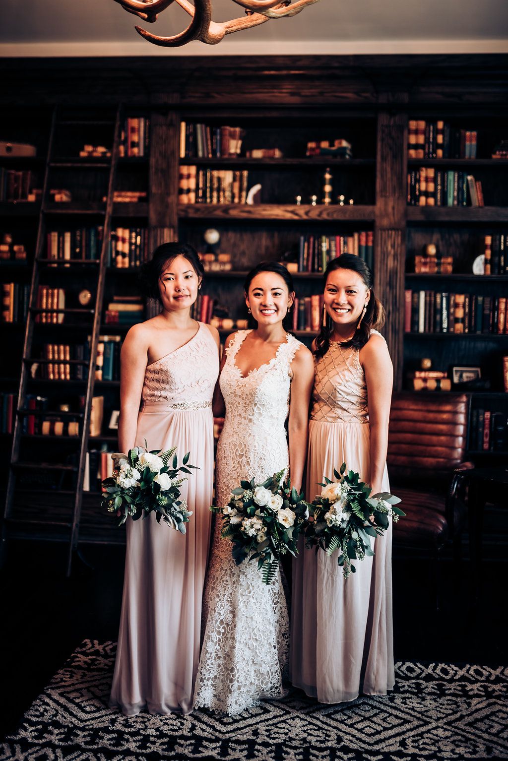 Three women in formal attire hold white floral bouquets while posing in front of library bookshelves.