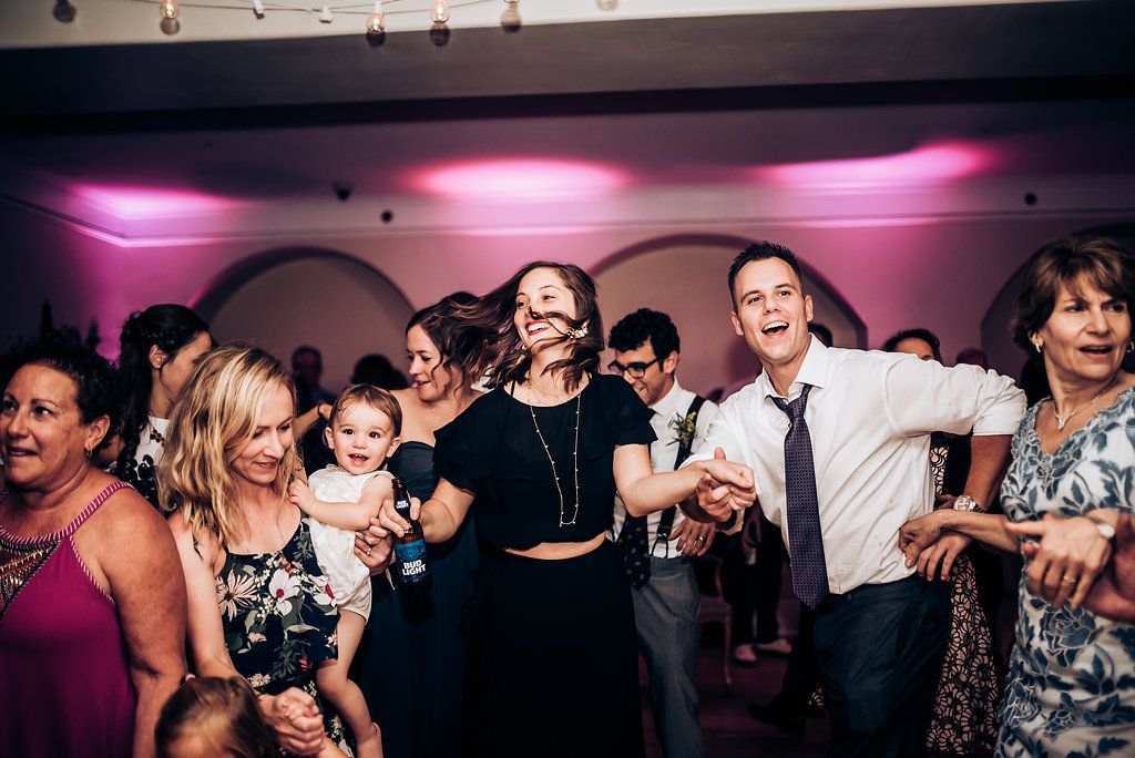 Guests dancing energetically at a wedding reception under pink mood lighting and string lights.