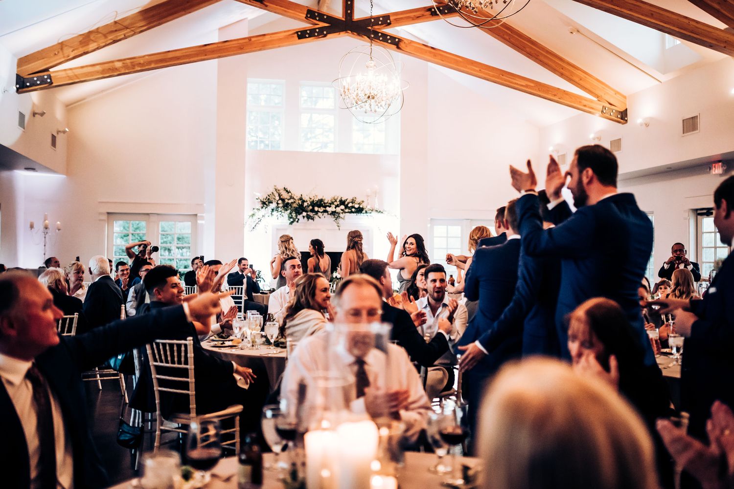 Wedding guests celebrating with raised hands in an elegant reception hall with exposed beams.