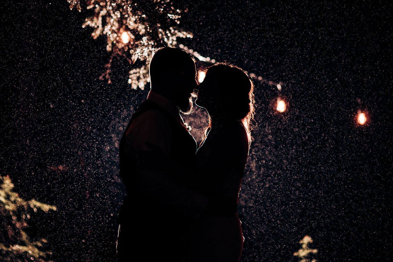 Silhouetted couple stands beneath string lights in evening rain.