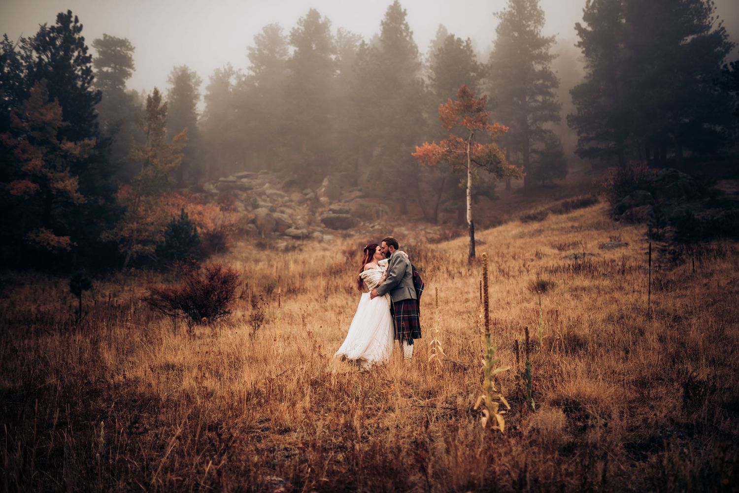 A wedding couple stands in a misty mountainside meadow with evergreen trees and golden grass in autumn.