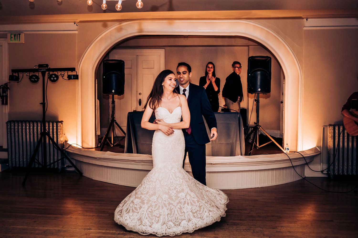A bride in a lace mermaid gown poses with her partner on a stage with an arched backdrop.