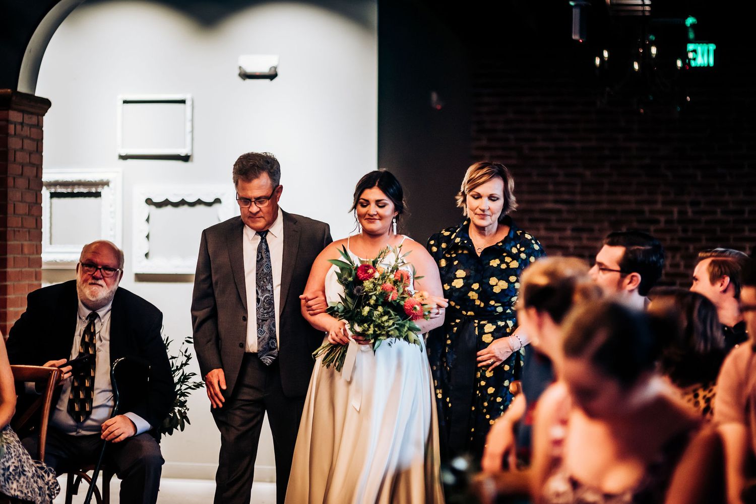 Wedding ceremony processional in an indoor venue with exposed brick walls and elegant chandeliers.