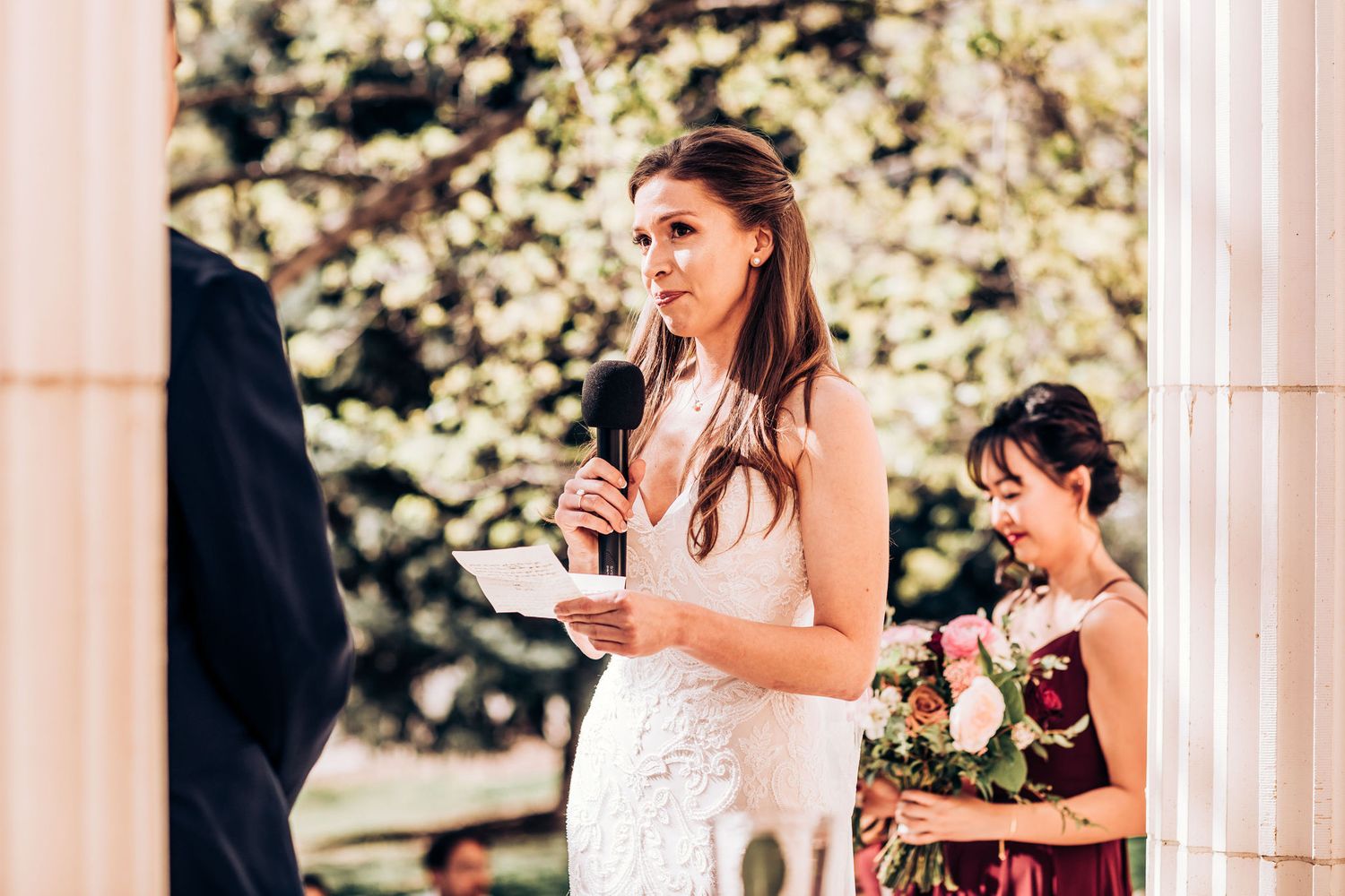 Wedding ceremony participants in formal attire standing between classical white columns outdoors.