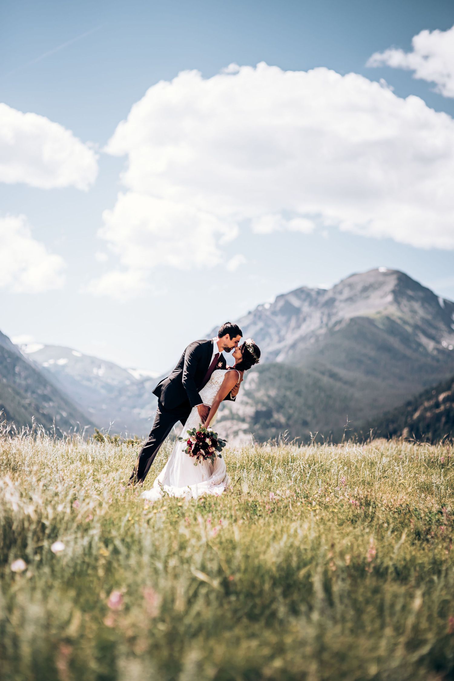 A romantic wedding couple embraces in a dramatic dip pose against a backdrop of majestic mountain peaks and wildflower fields.