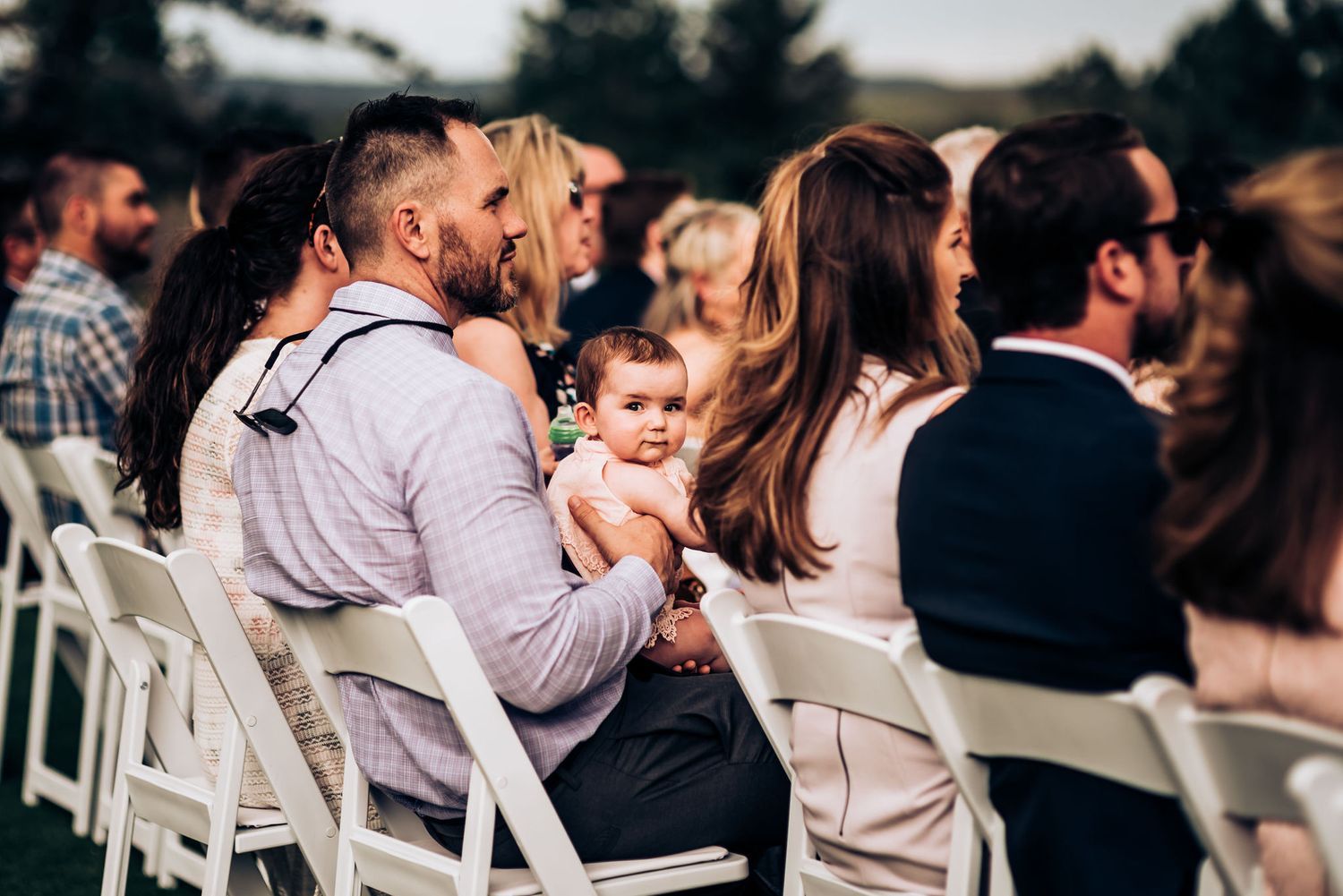 Wedding guests seated in white chairs during an outdoor ceremony.