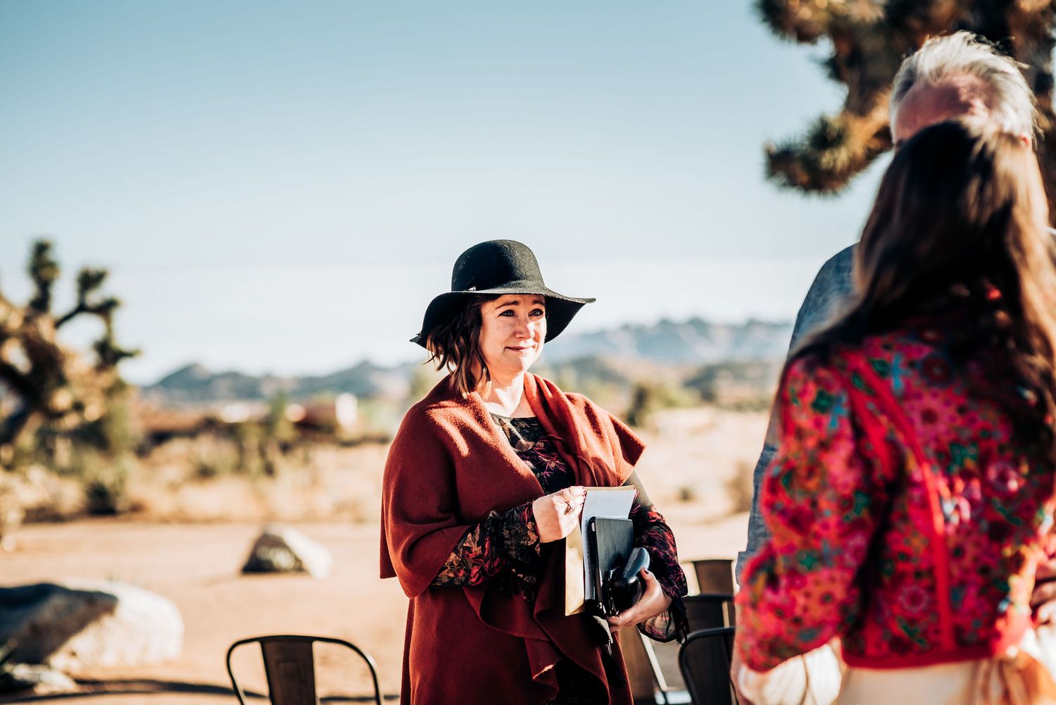 Guest in burgundy dress and black hat pours drinks at desert wedding reception.